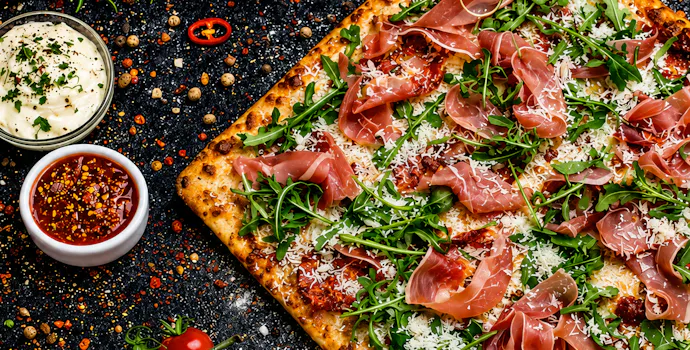 Pizza cu prosciutto, rucola și brânză, înconjurată de sosuri, roșii cherry și condimente pe un fundal artistic.