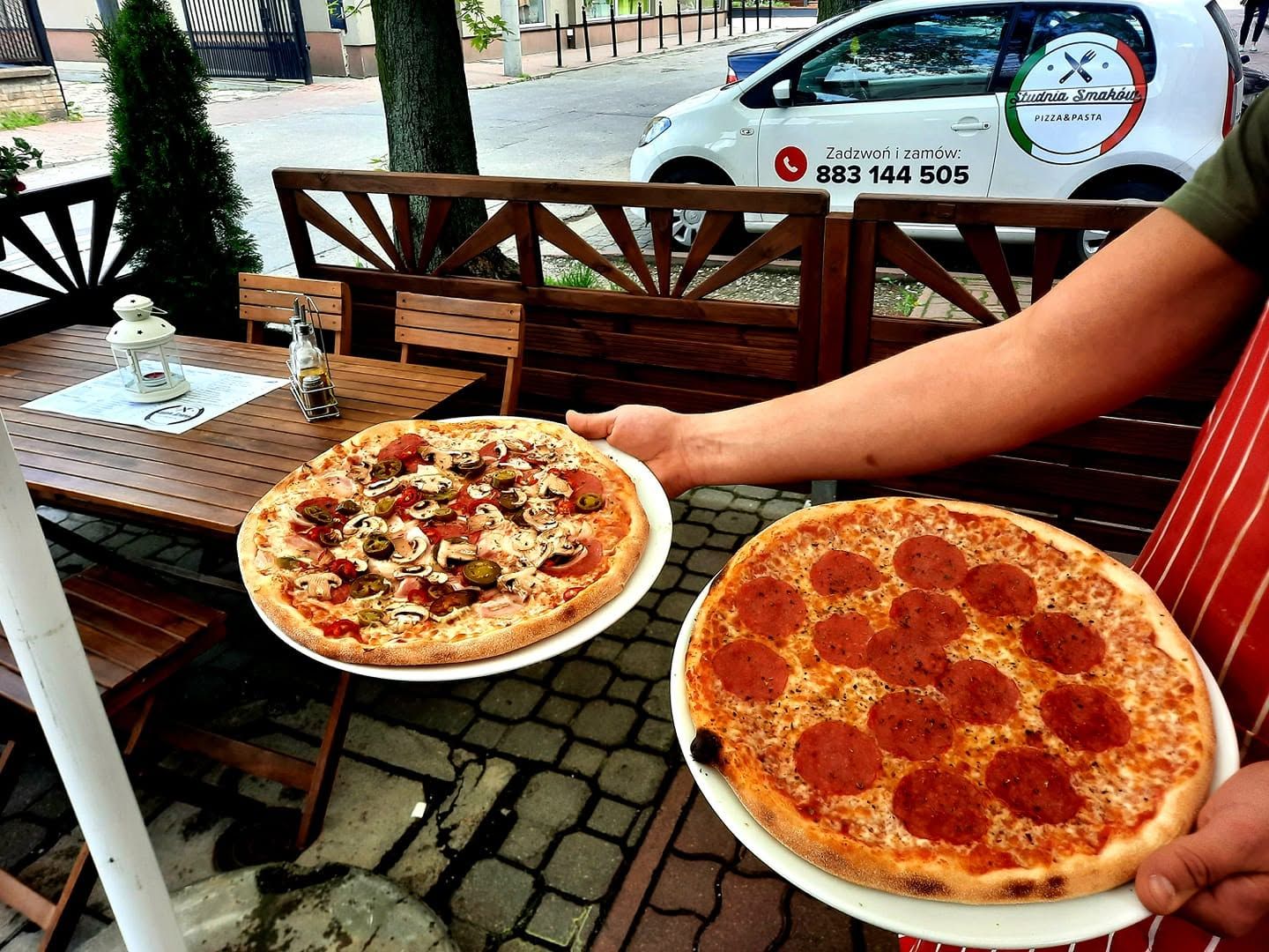 Osoba trzymająca dwa pizze: jedna z pieczarkami, druga z pepperoni, na tle restauracyjnego tarasu.