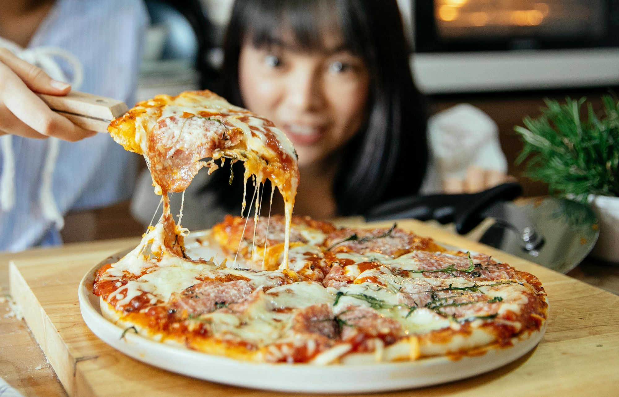 Kobieta trzyma kawałek serowej pizzy, na tle blatu kuchennego i piekarnika.