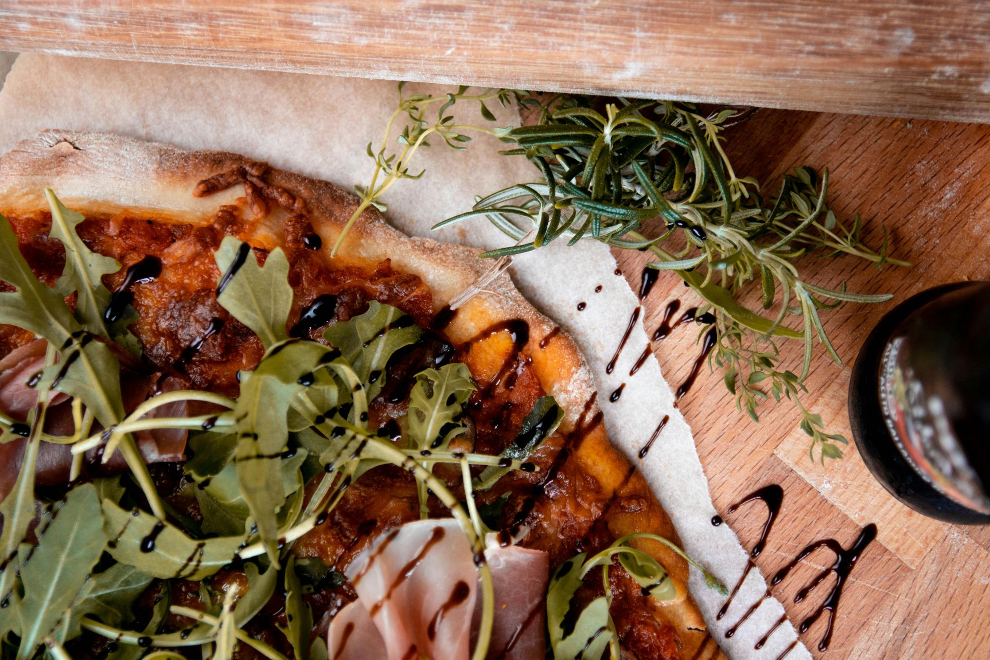 Pizza z szynką, rukolą i ziołami, polana sosem balsamicznym, na drewnianym blacie kuchennym.