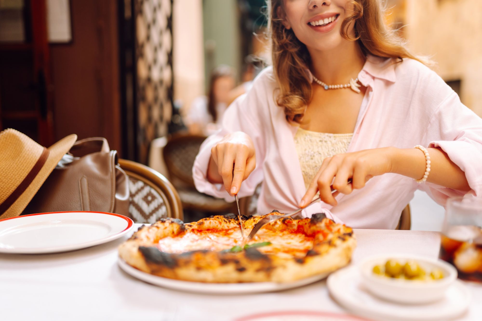 Młoda kobieta kroi pizzę przy stole w restauracji, wokół talerze i oliwki.