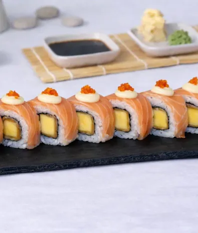 Rulouri sushi cu somon, mango și icre, servite pe un platou de piatră, alături de sos de soia și condimente.