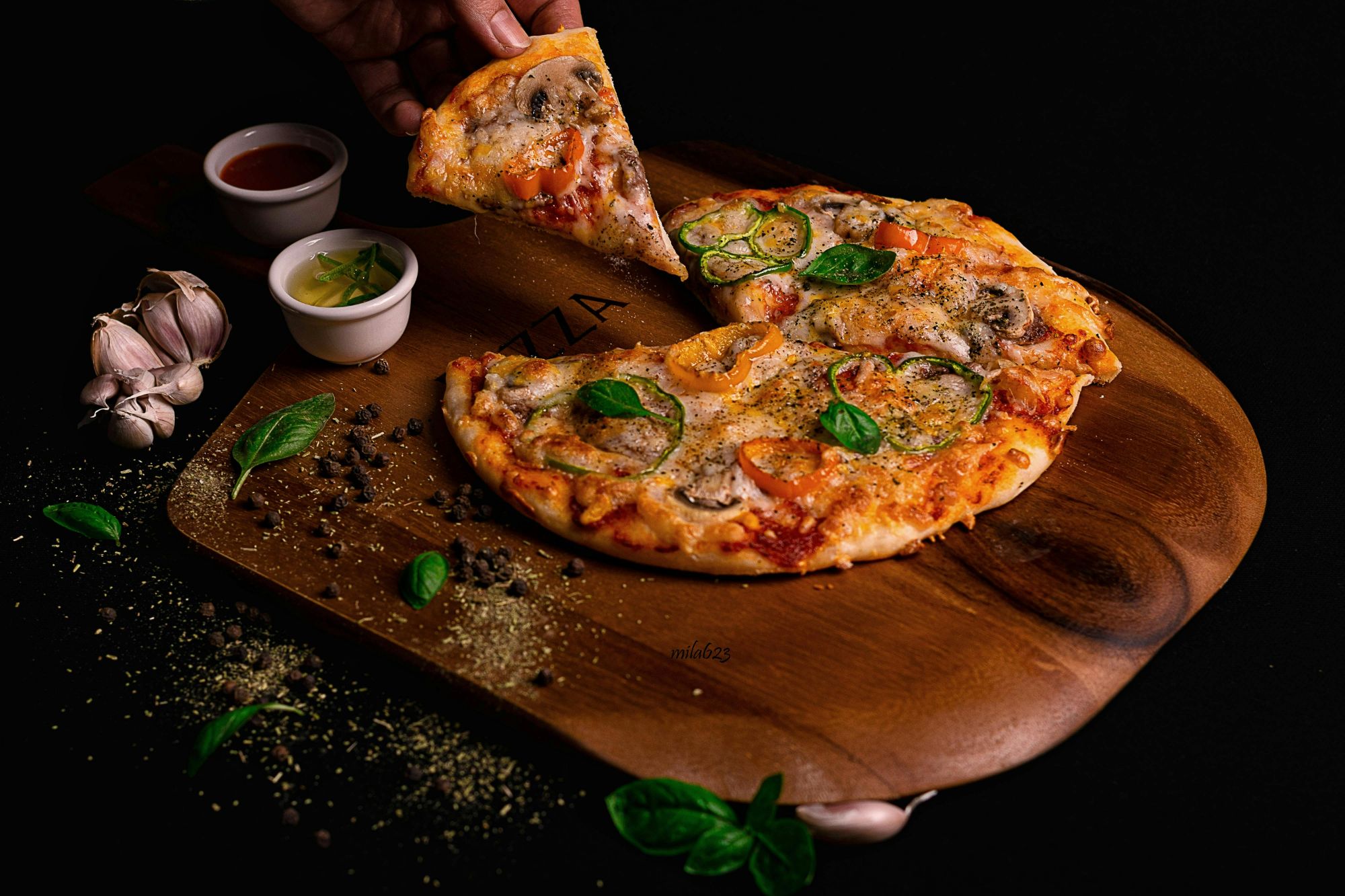 Pizza na drewnianej desce, kawałek wyjmowany, obok sosy, czosnek i świeże zioła.