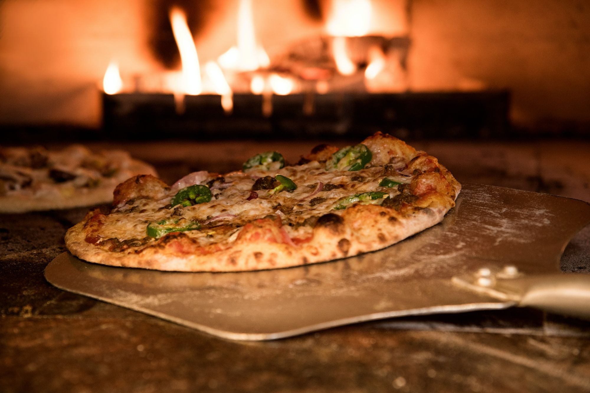 Pyszna pizza z pieca opalanego drewnem, z jalapeño, na łopacie do pizzy w tle płonący ogień.
