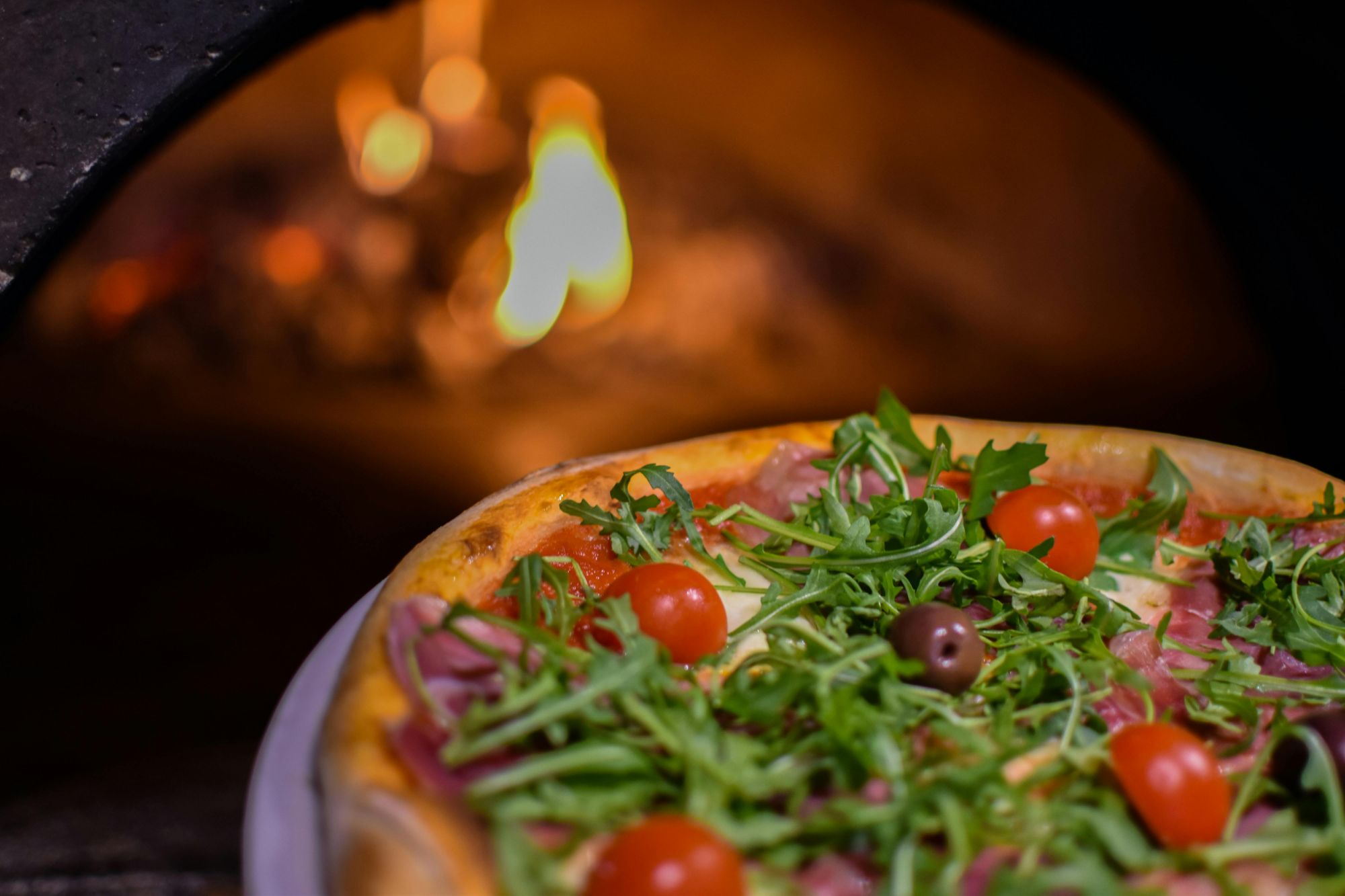 Pyszna pizza z szynką, rukolą, pomidorkami i oliwkami, pieczona w piecu opalanym ogniem.