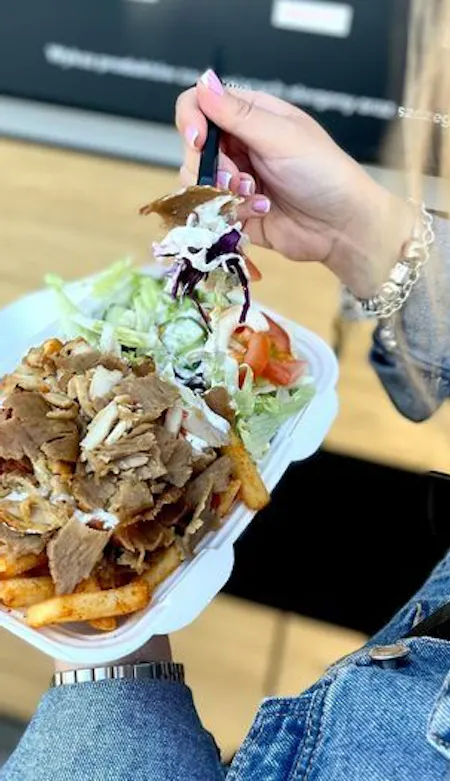 Osoba trzymająca talerz z frytkami, sałatą i mięsem kebab, w tle widoczna ciemna ściana.
