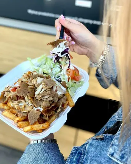 Osoba trzymająca talerz z frytkami, sałatą i mięsem kebab, w tle widoczna ciemna ściana.