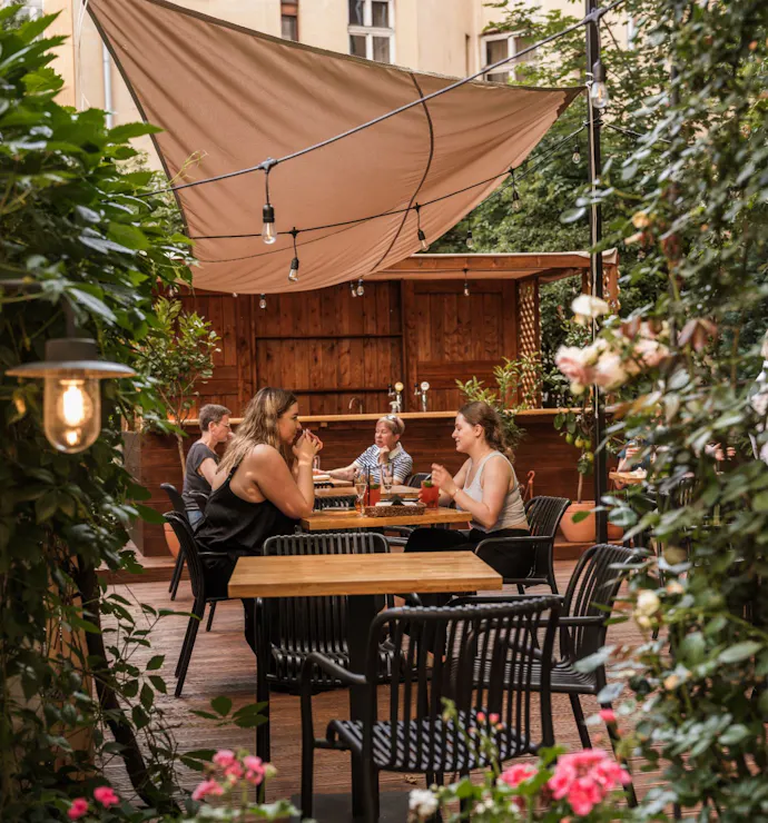Ludzie siedzący przy stole w ogrodzie restauracyjnym otoczonym roślinnością i kwiatami.