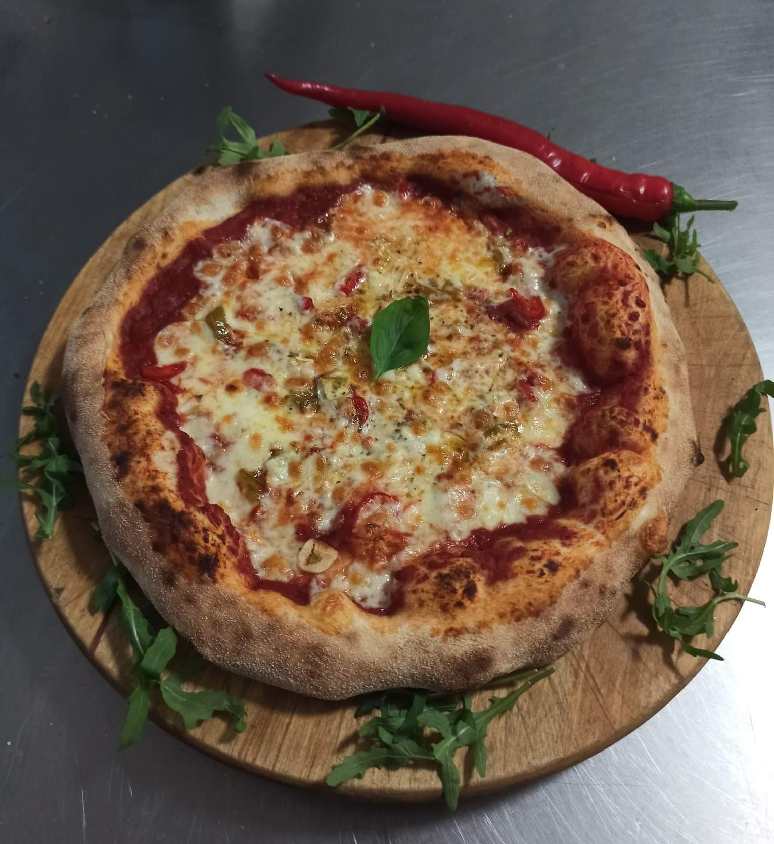 Pizza na drewnianej desce, udekorowana bazylią, papryczką chili i świeżymi ziołami.