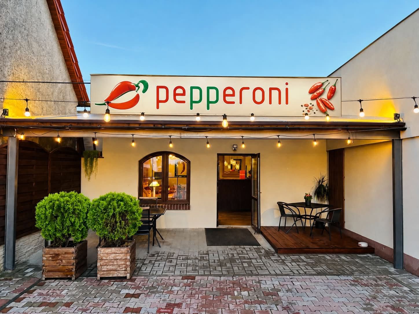 Restauracja Pepperoni z widocznym znakiem i oświetleniem, otoczenie z roślinami i meblami na zewnątrz.