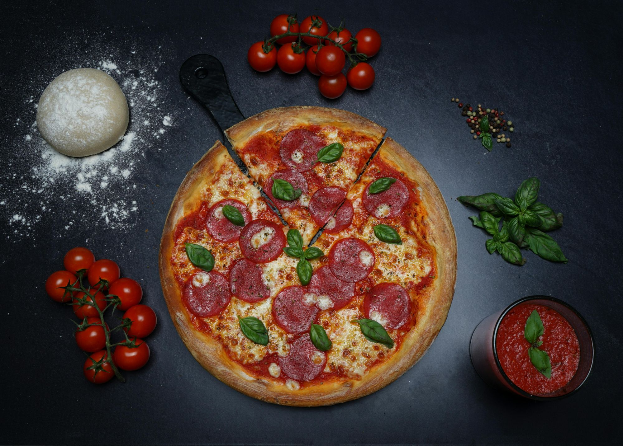 Pizza pepperoni na czarnym tle, obok świeże pomidory, bazyliowe liście i sos pomidorowy.