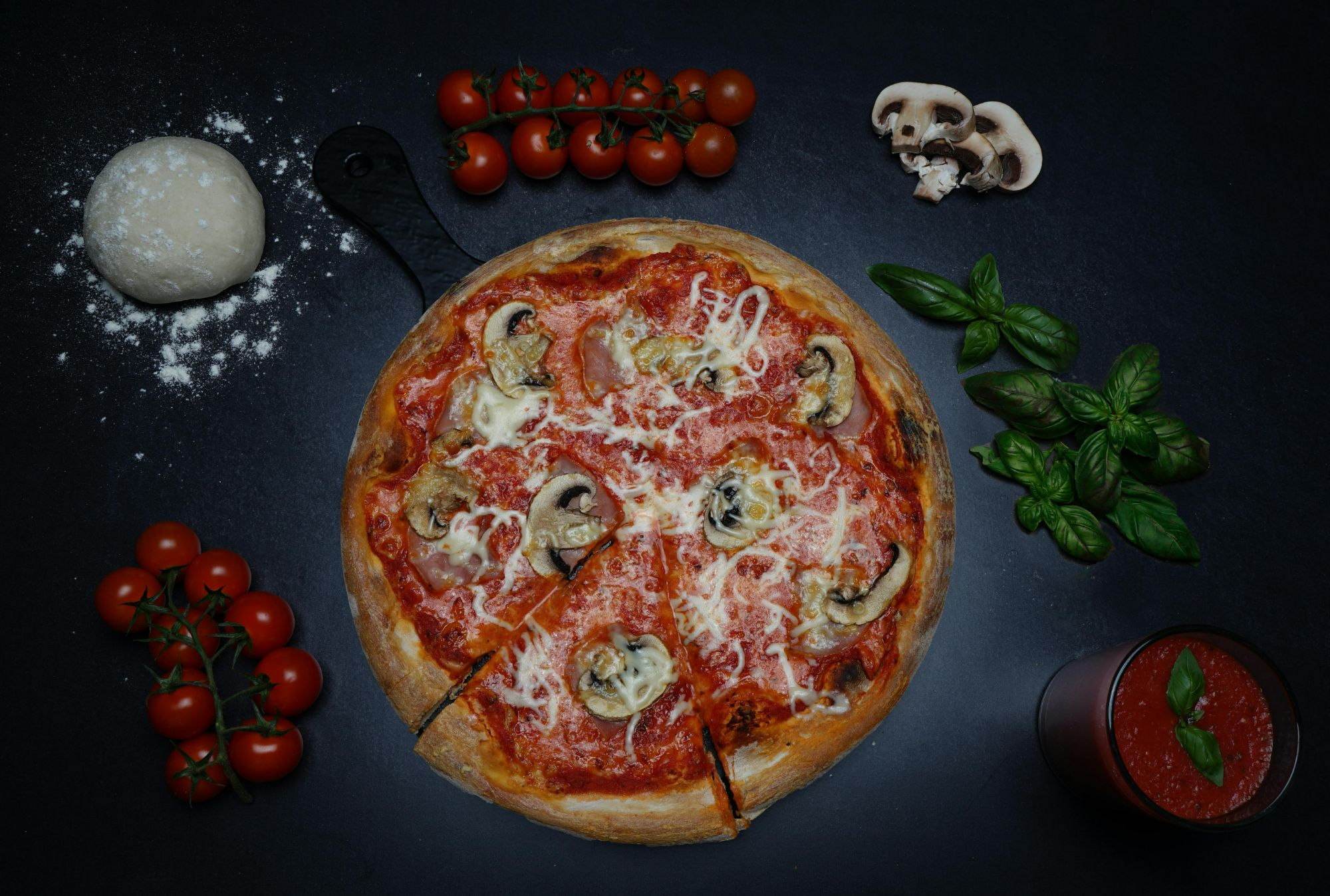 Pizza z sosem pomidorowym, serem, pieczarkami i bazylią, otoczona pomidorami, ciastem i kubkiem sosu.