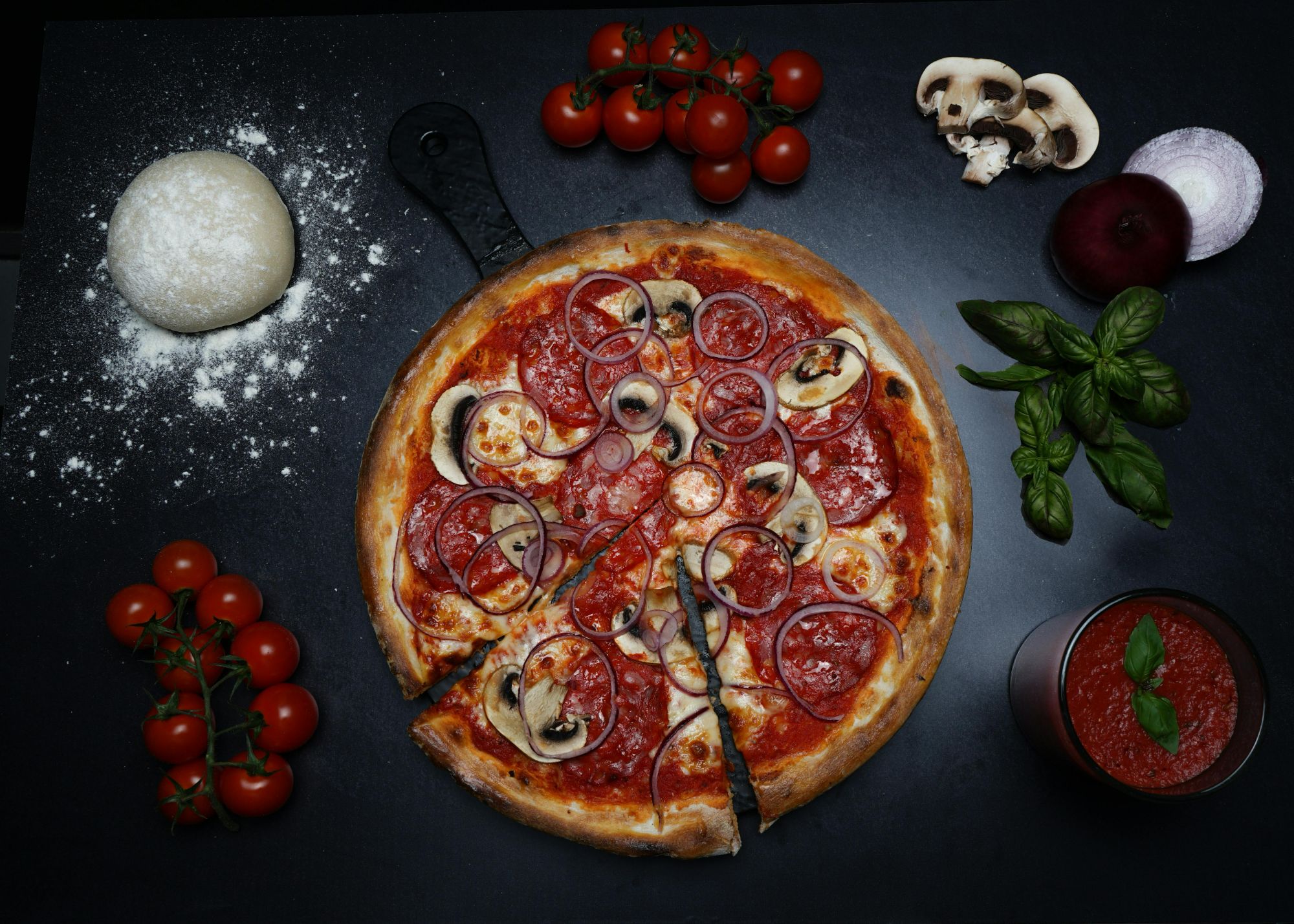 Pizza z pepperoni, serem, pieczarkami i cebulą, otoczona świeżymi składnikami na czarnym tle.
