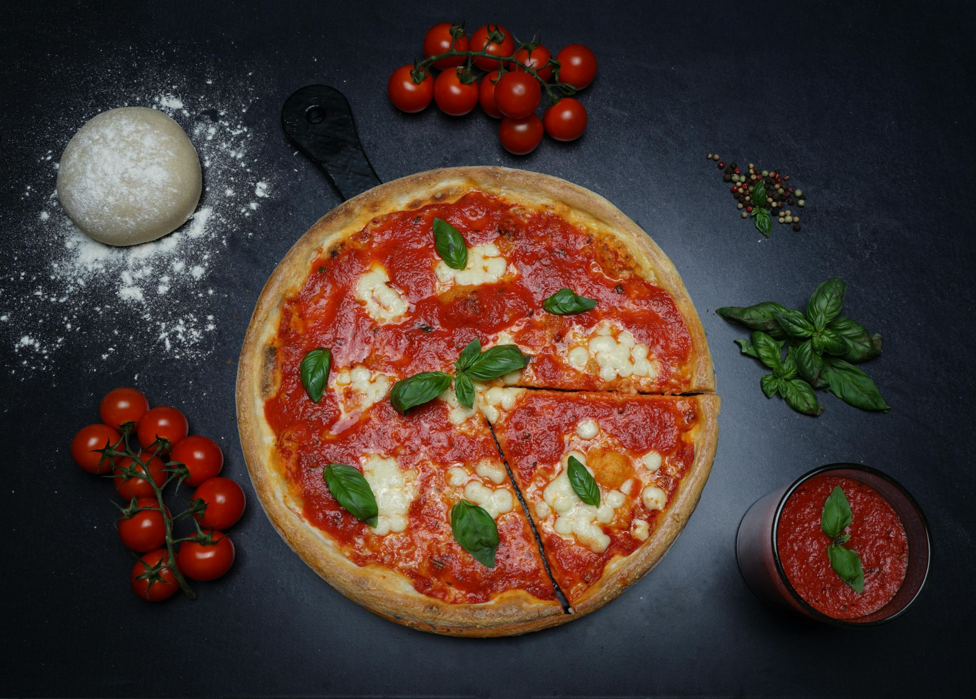 Pizza Margherita na czarnym tle, otoczona pomidorami, bazylią, mąką i sosem pomidorowym.