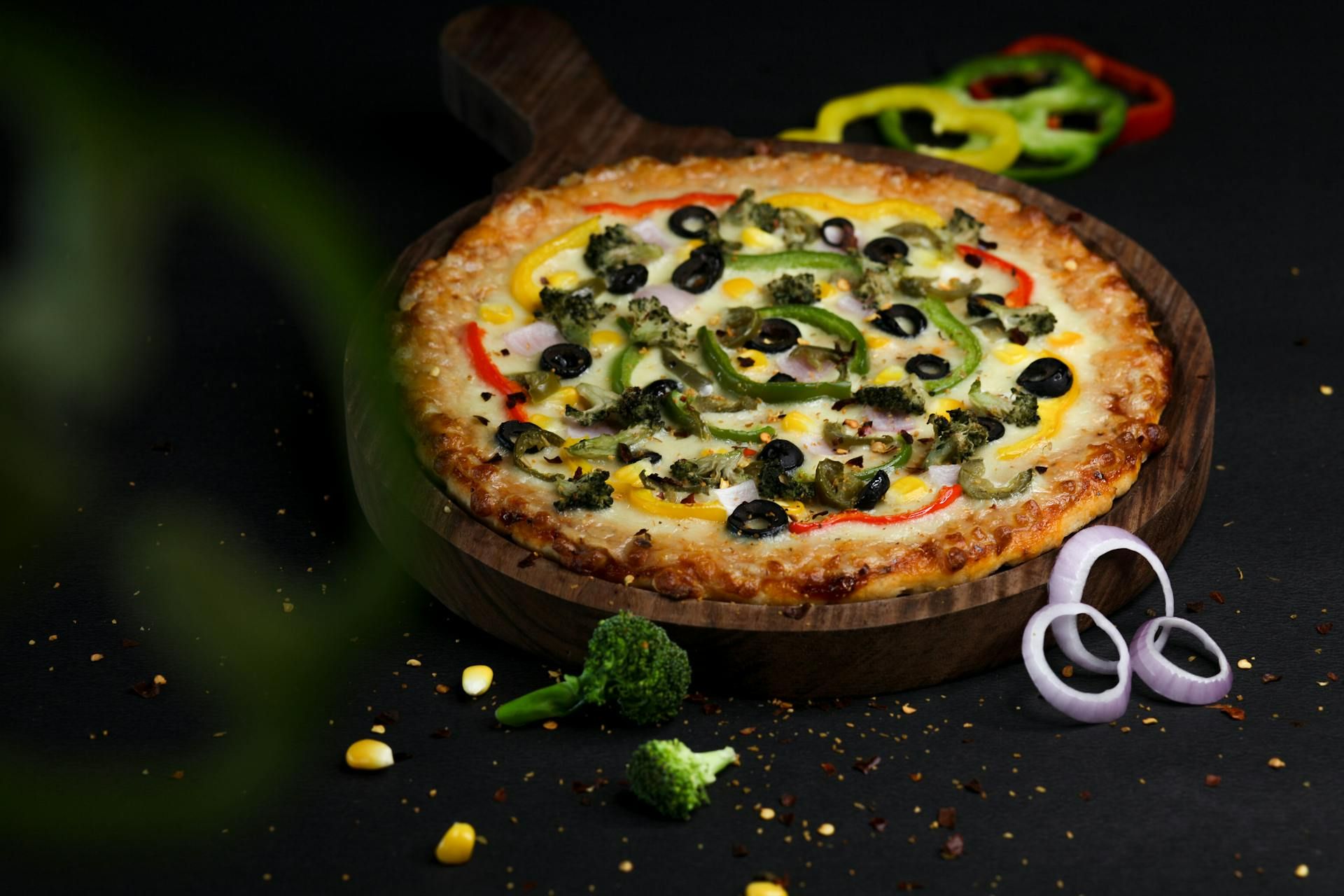 Pizza na drewnianej desce, z serem, oliwkami, papryką i cebulą, na czarnym tle.
