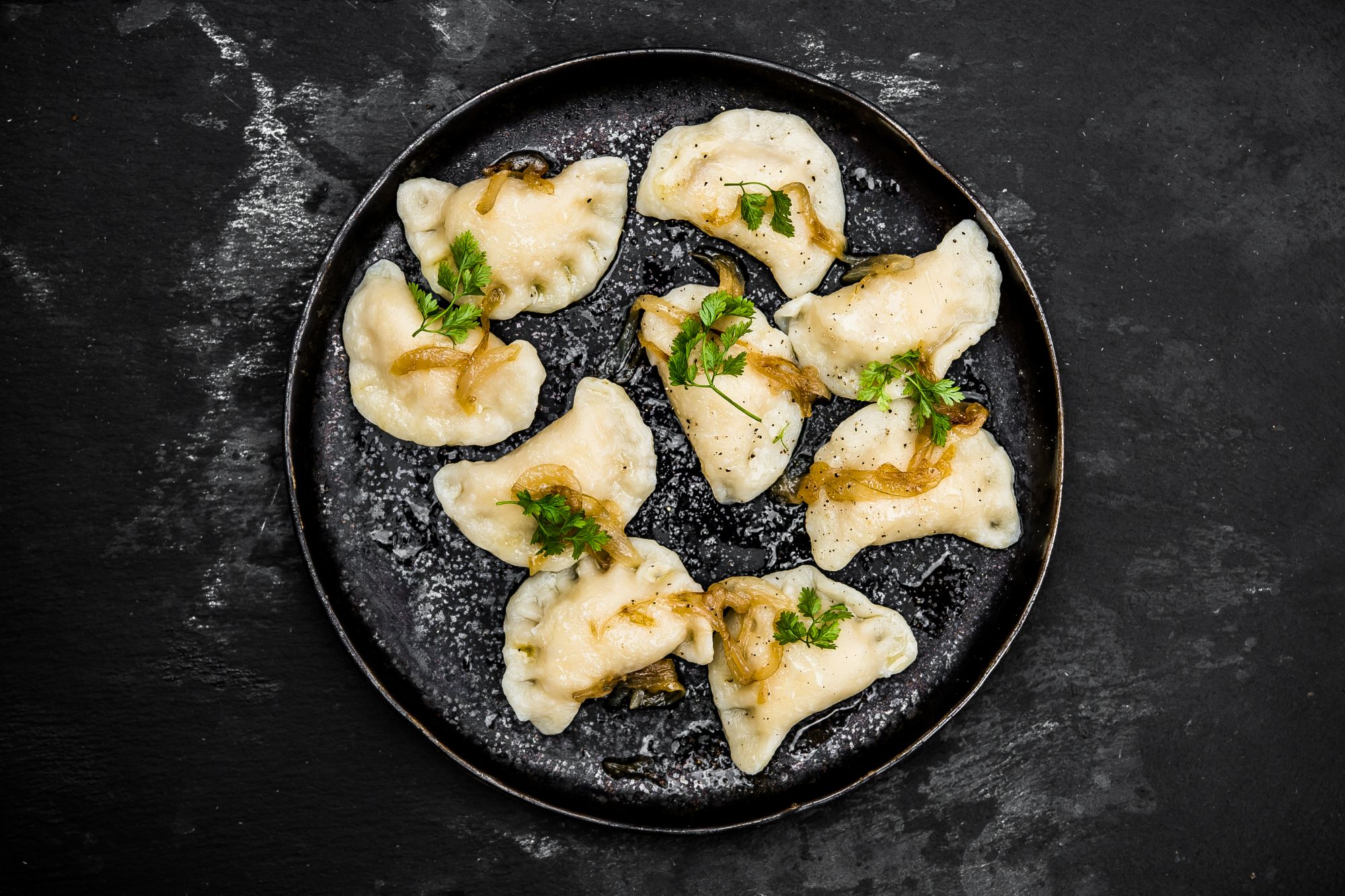 Pierogi na czarnym talerzu, polane cebulą i posypane świeżą pietruszką.