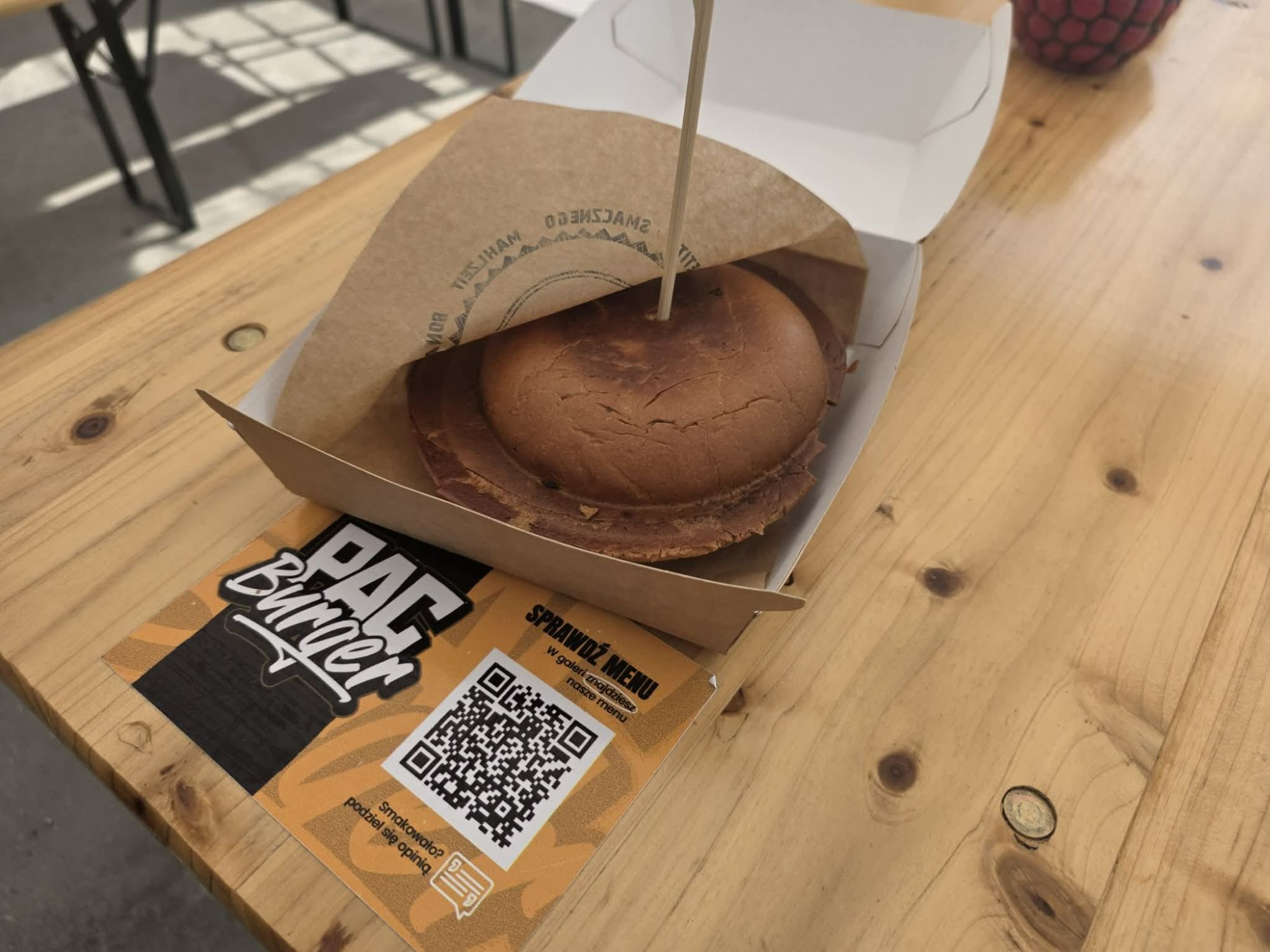 Burger w opakowaniu na drewnianym stole, obok karta z kodem QR i nazwą lokalu.