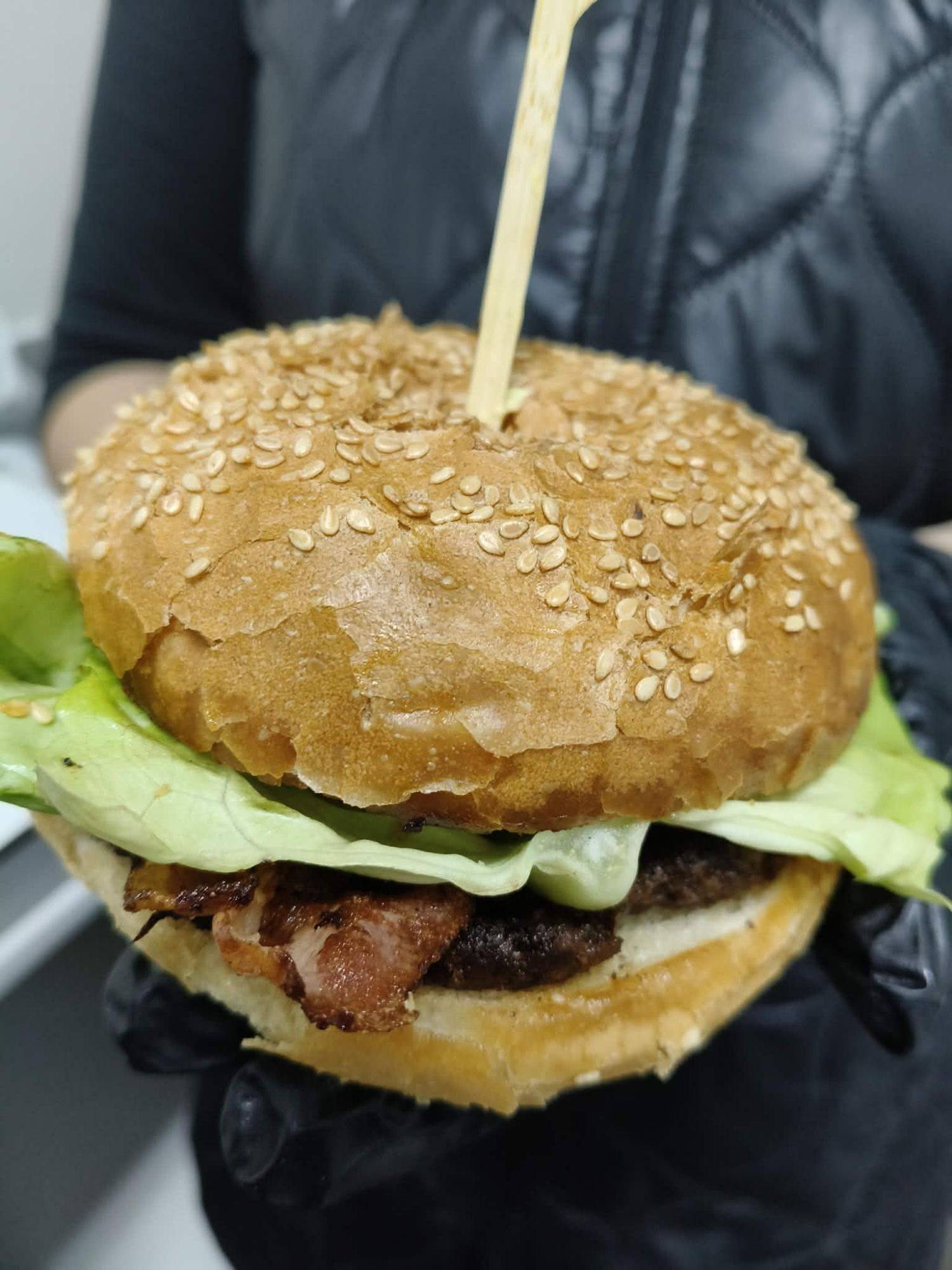 Zbliżenie na burgera z serem, bekonem i sałatą w sezamowej bułce trzymanej w ręce.