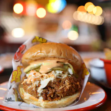 Soczysty burger z kurczakiem, sałatą i sosami, podany na talerzu w tle z rozmytymi światłami.