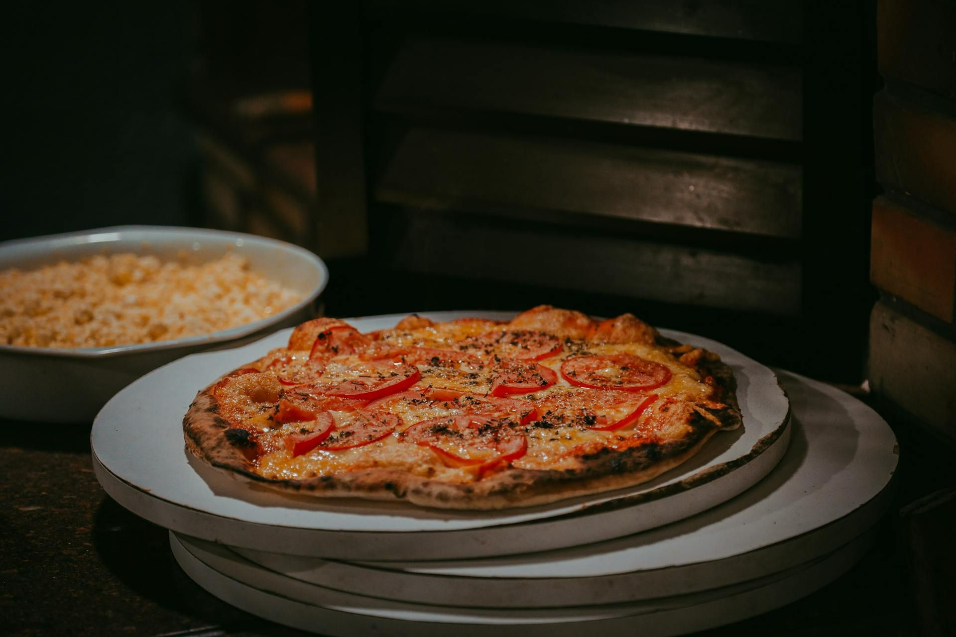 Pizza z serem, pomidorami i przyprawami na desce, obok talerz z makaronem.