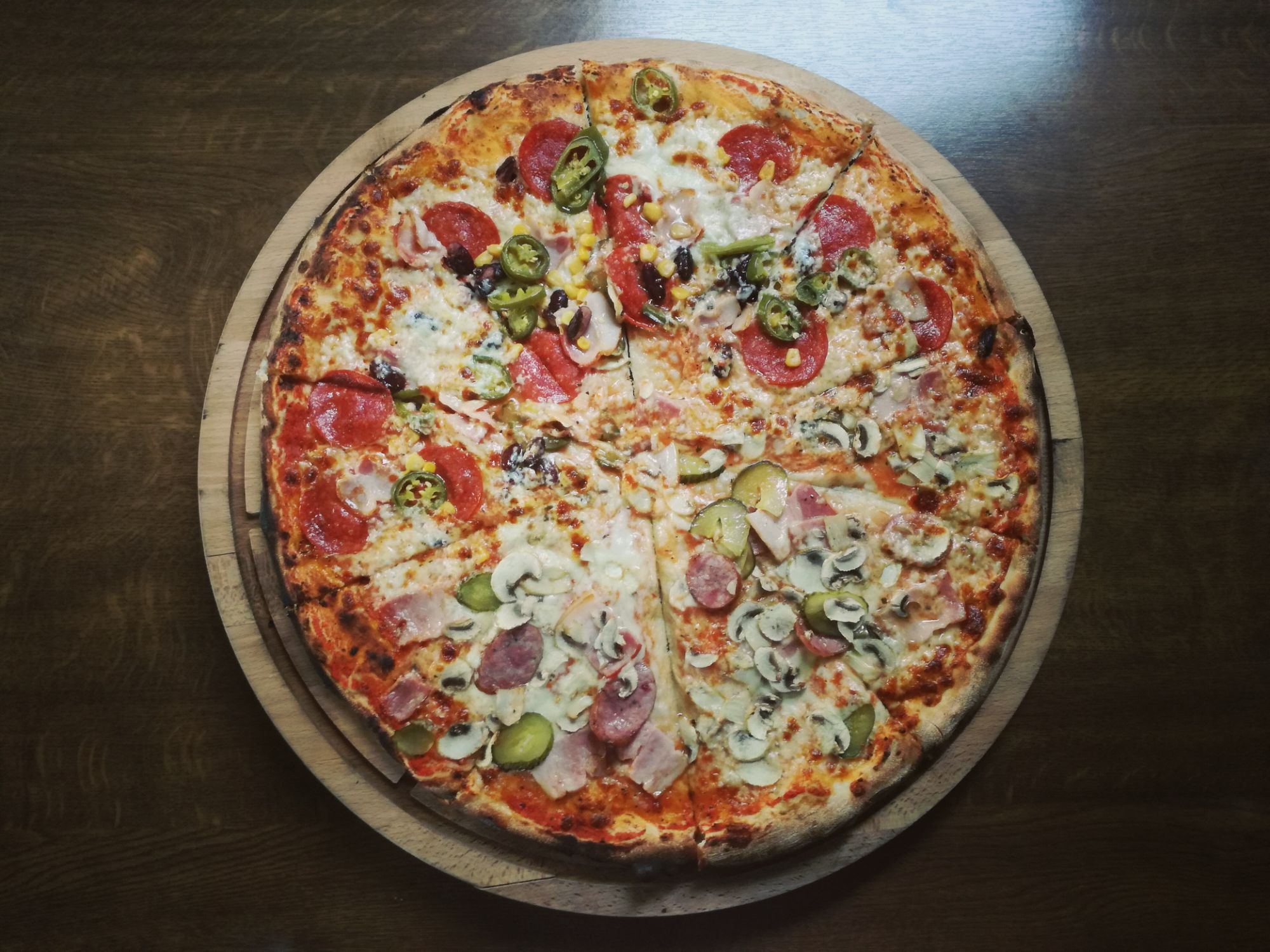 Pizza z różnymi składnikami, w tym pepperoni, pieczarkami i jalapeño, na desce drewnianej.