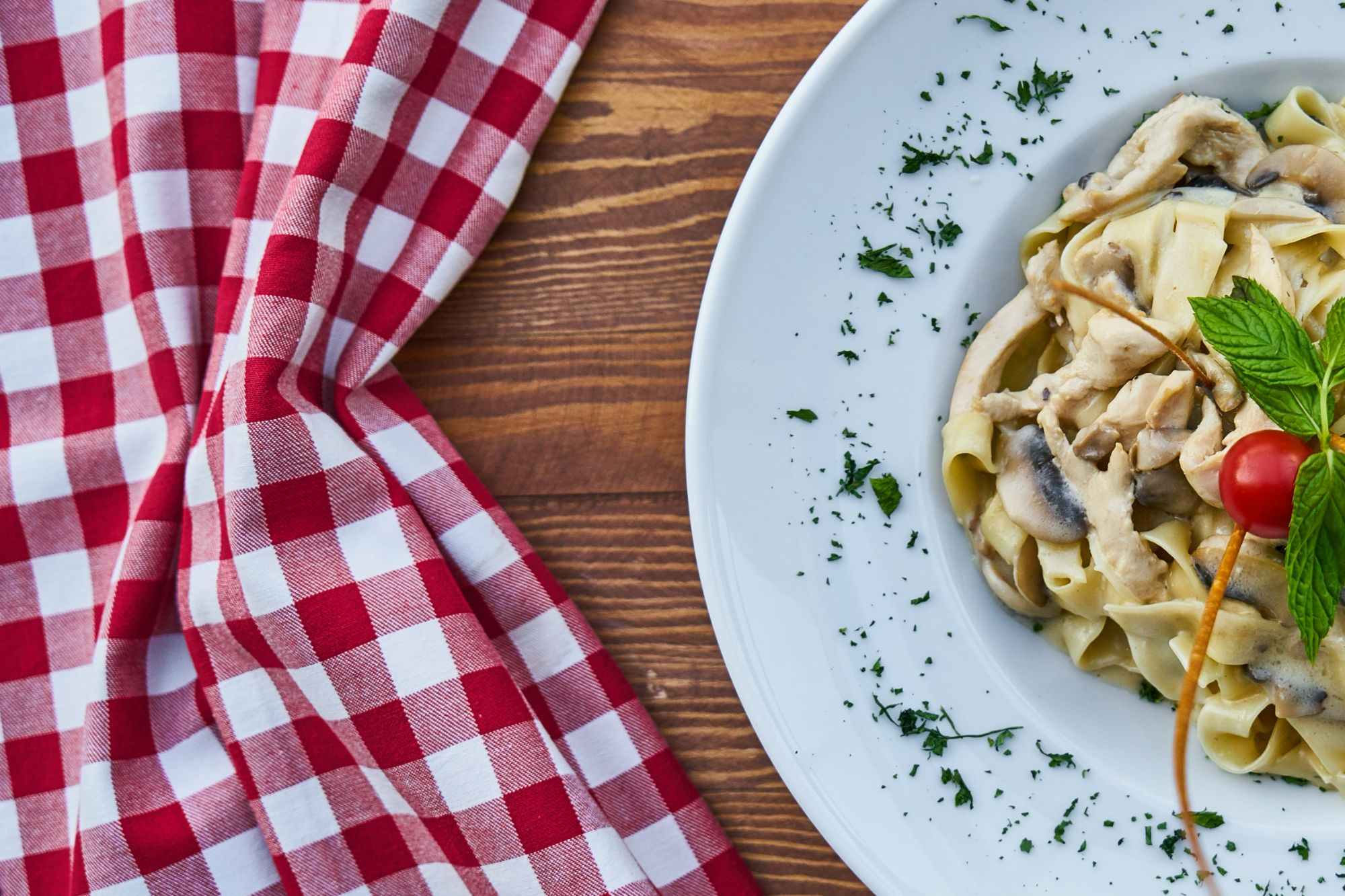 Makaron fettuccine z sosem grzybowym, pomidorem koktajlowym i świeżą miętą na tle czerwono-białej kratki.