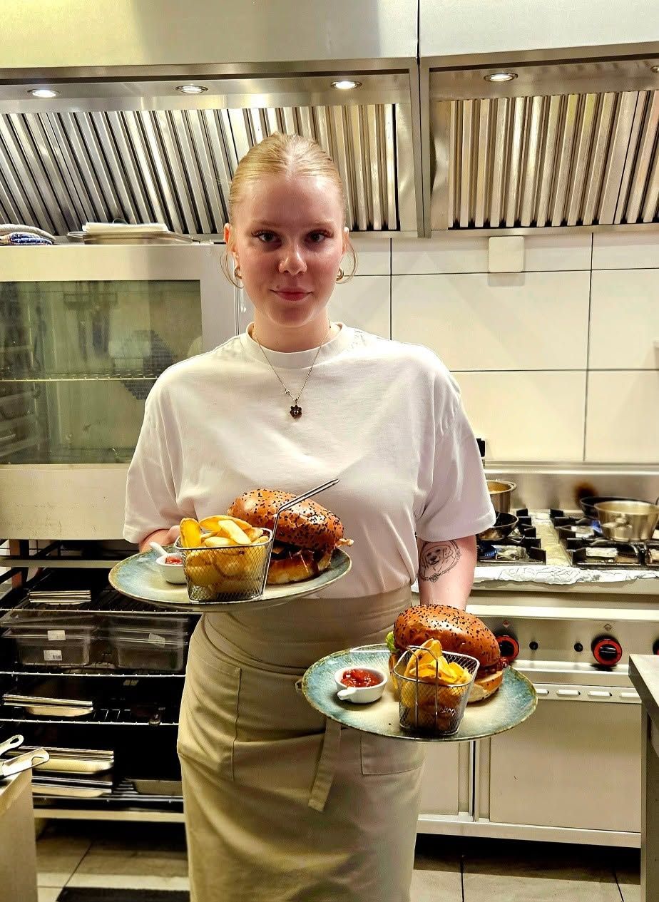 Kobieta w kuchni trzymająca talerze z hamburgerami i frytkami, w tle sprzęt gastronomiczny.