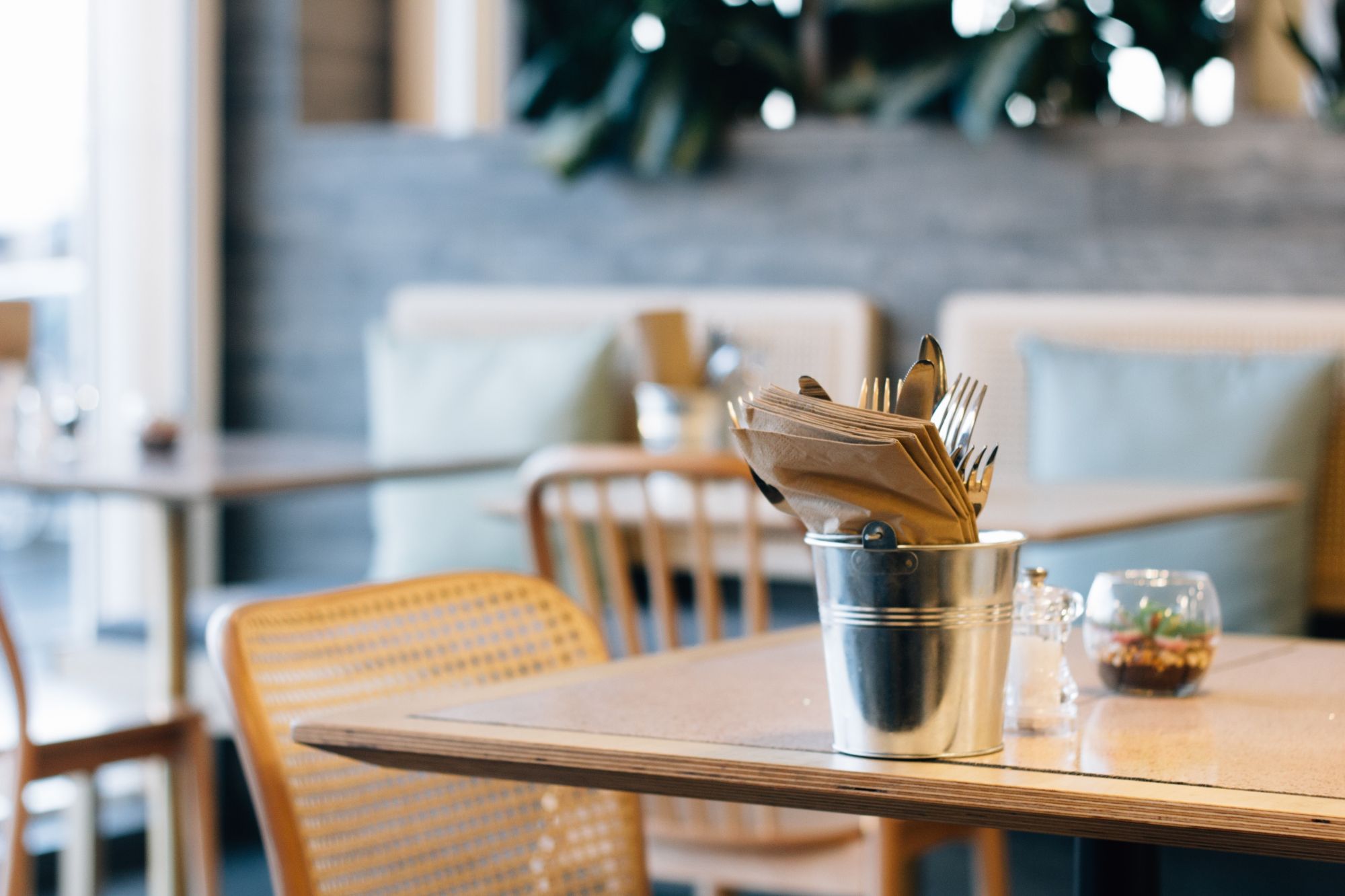 Stół w restauracji z metalowym pojemnikiem na sztućce, drewnianymi krzesłami i roślinnymi dekoracjami.