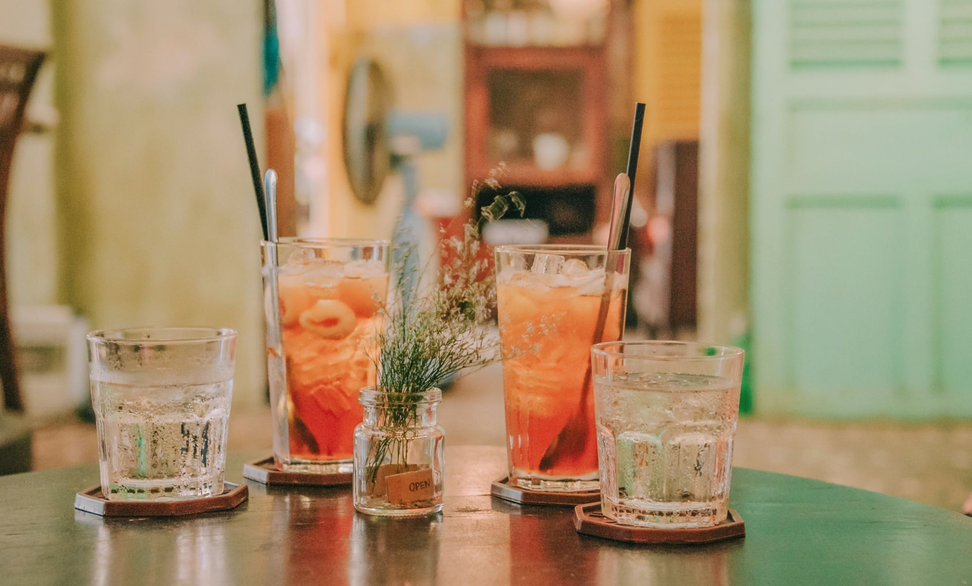 Trzy szklanki z drinkami i jedna z wodą, ozdobione małą rośliną na stole w stylowym wnętrzu.