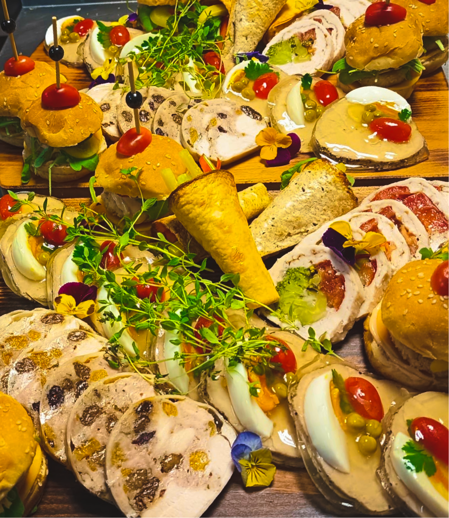Różnorodne przekąski: mini burgery, wędliny, sałatki, oraz desery na drewnianej tacy, udekorowane ziołami i kwiatami.