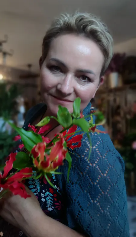 Kobieta z krótkimi włosami trzyma kolorowe kwiaty, uśmiechając się w otoczeniu roślin w jasnym wnętrzu.