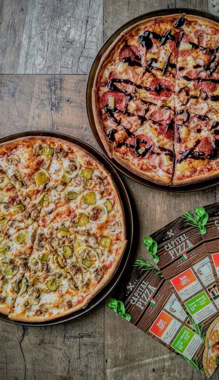 Dwie pizze na okrągłych talerzach: jedna z pieczarkami i jalapeño, druga z boczkiem i sosem. Obok ulotki pizzerii.
