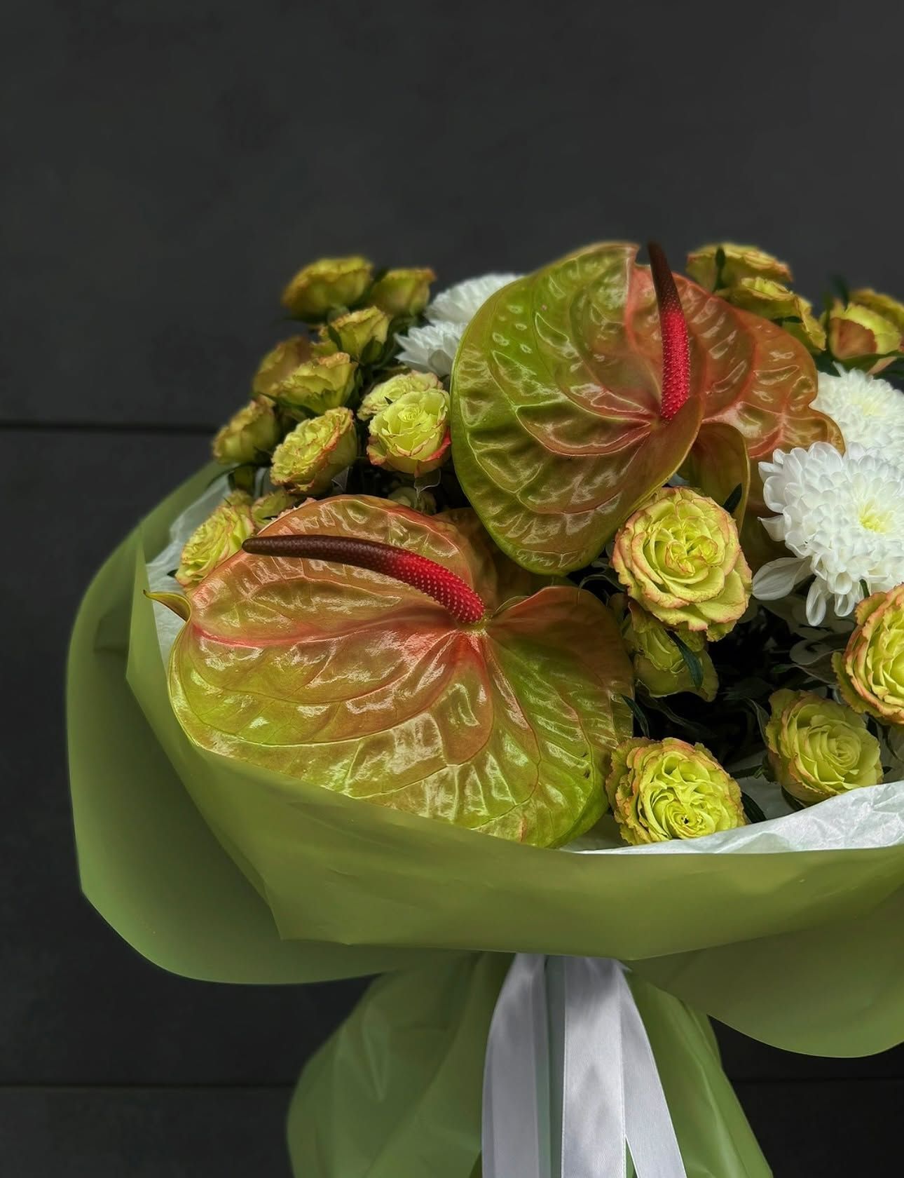 Bukiet kwiatów z anthurium, różami i chryzantemami na ciemnym tle, ozdobiony zieloną wstążką.