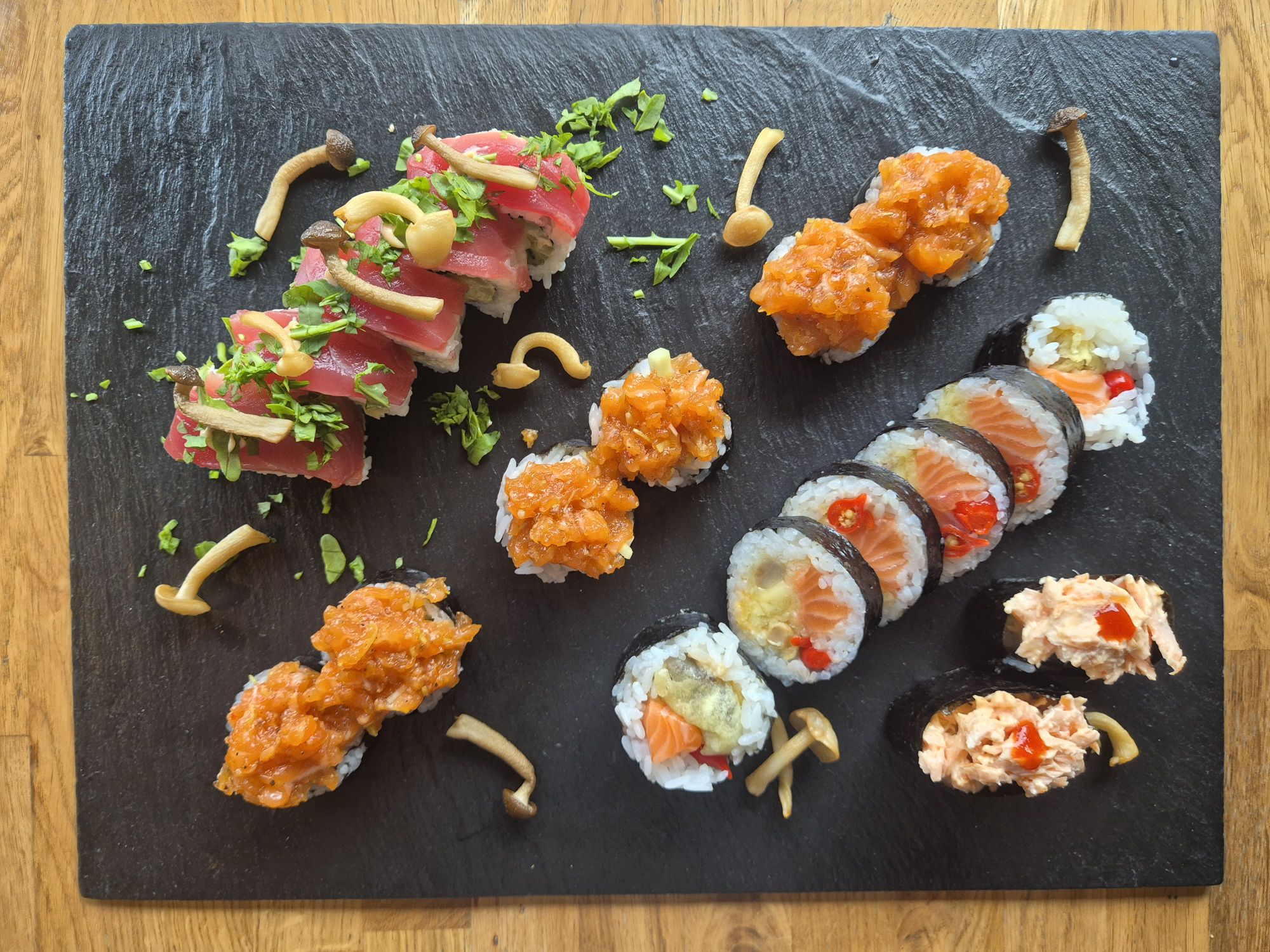 Deska z sushi z różnymi rolkami, w tym tuńczykiem, łososiem i warzywami, ozdobiona ziołami i grzybami.