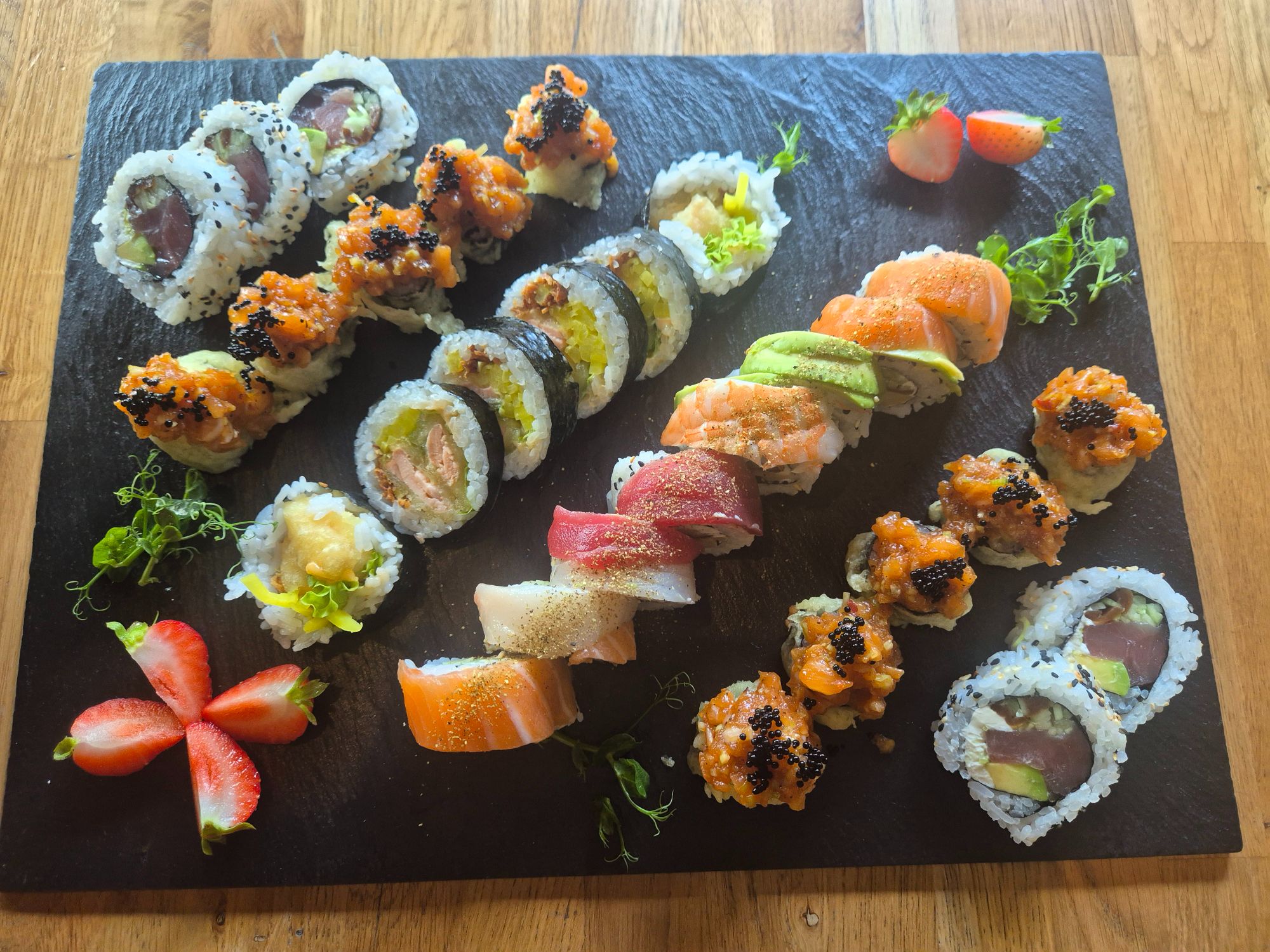 Sushi na czarnym talerzu, różnorodne rolki z rybami, warzywami i owocami, ułożone dekoracyjnie.