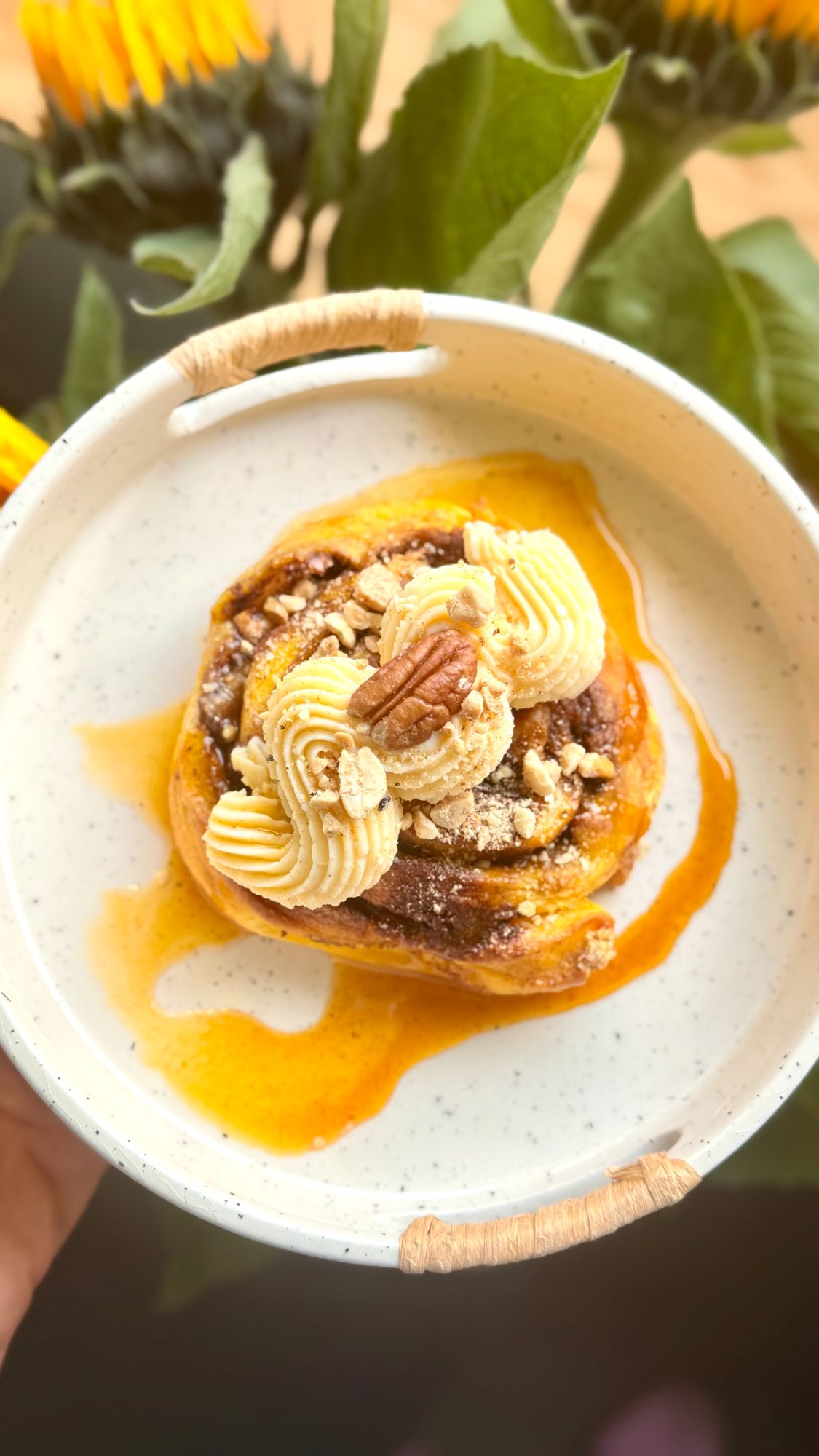 Cinnamon yeast roll with cream, nuts, and syrup against a backdrop of sunflowers.