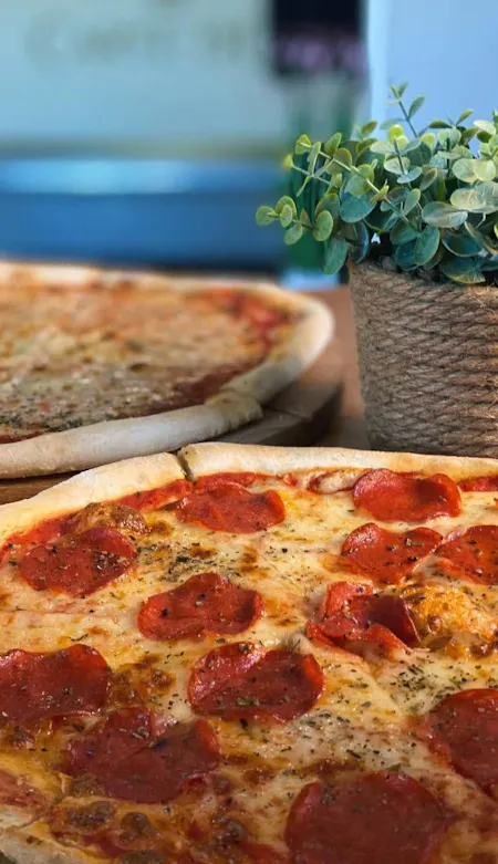 Pizza z pepperoni na drewnianym talerzu obok doniczki z rośliną, w tle druga pizza.