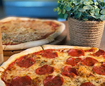 Pizza z pepperoni na drewnianym talerzu obok doniczki z rośliną, w tle druga pizza.