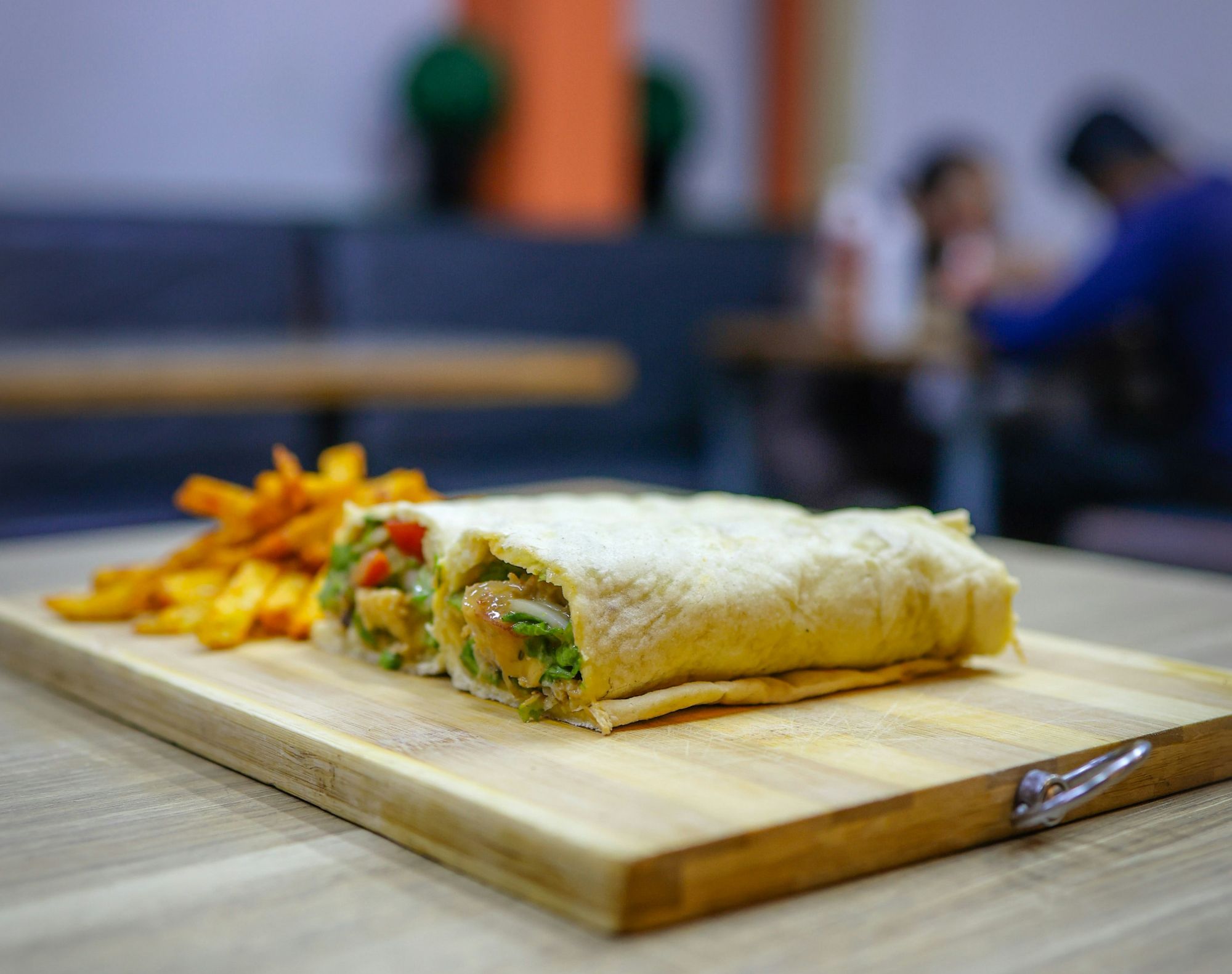Tortilla z nadzieniem warzywno-mięsnym na drewnianej desce, obok frytki, w tle kilka osób siedzących przy stole.