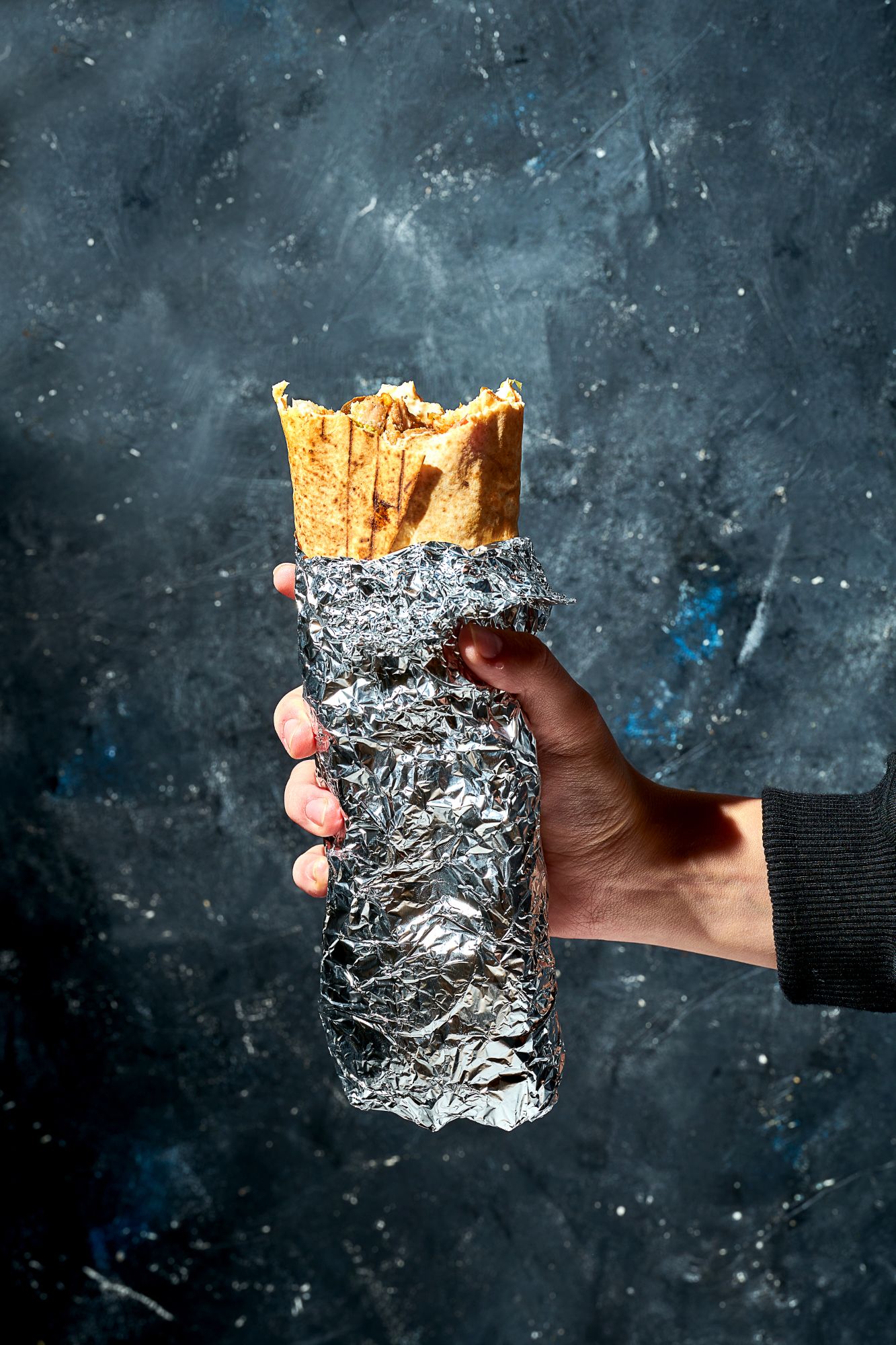 Ręka trzymająca burrito owinięte w folię aluminiową na ciemnym tle.