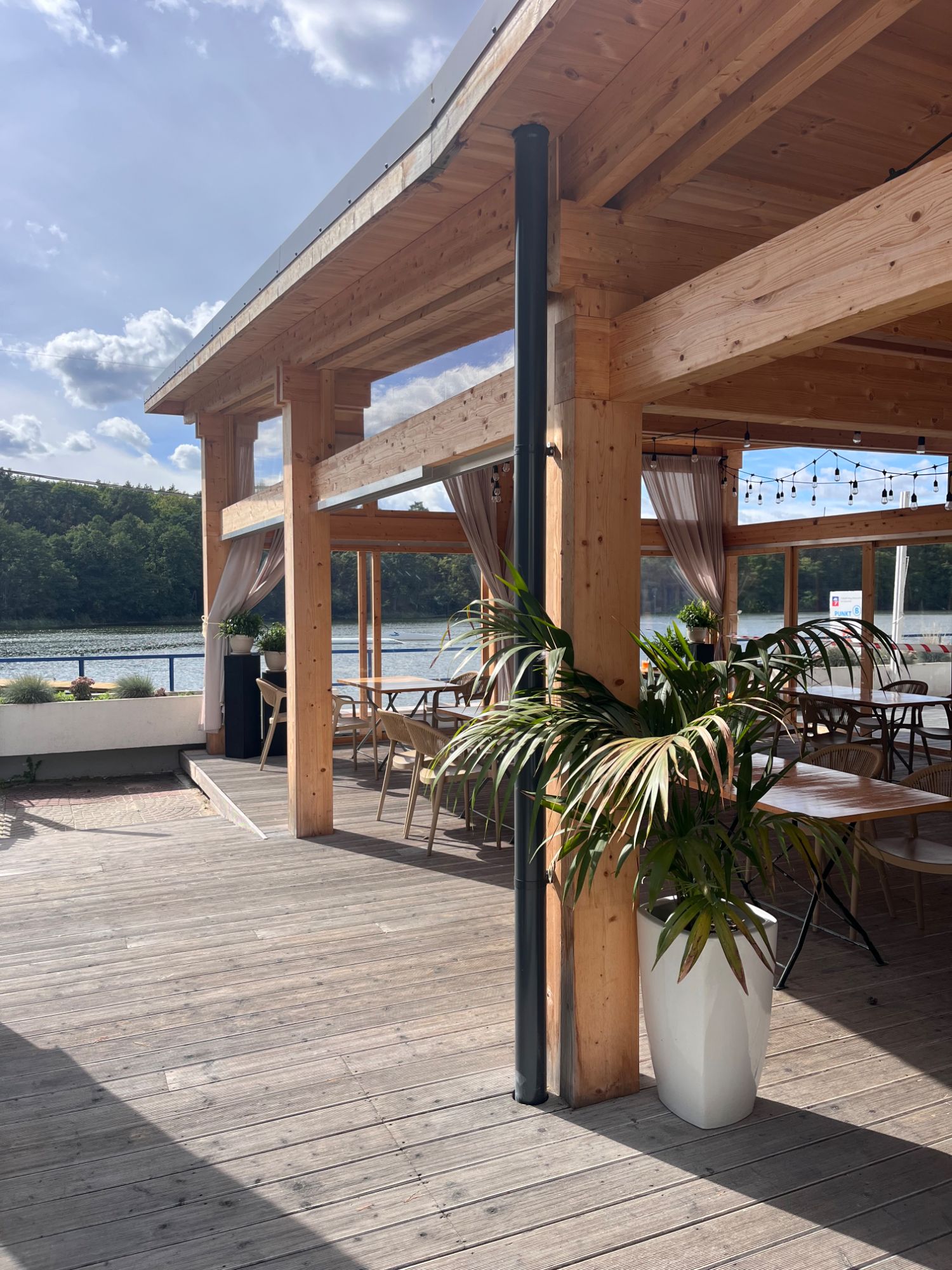 Nowoczesna restauracja przy wodzie z drewnianą konstrukcją, roślinnością i widokiem na jezioro.