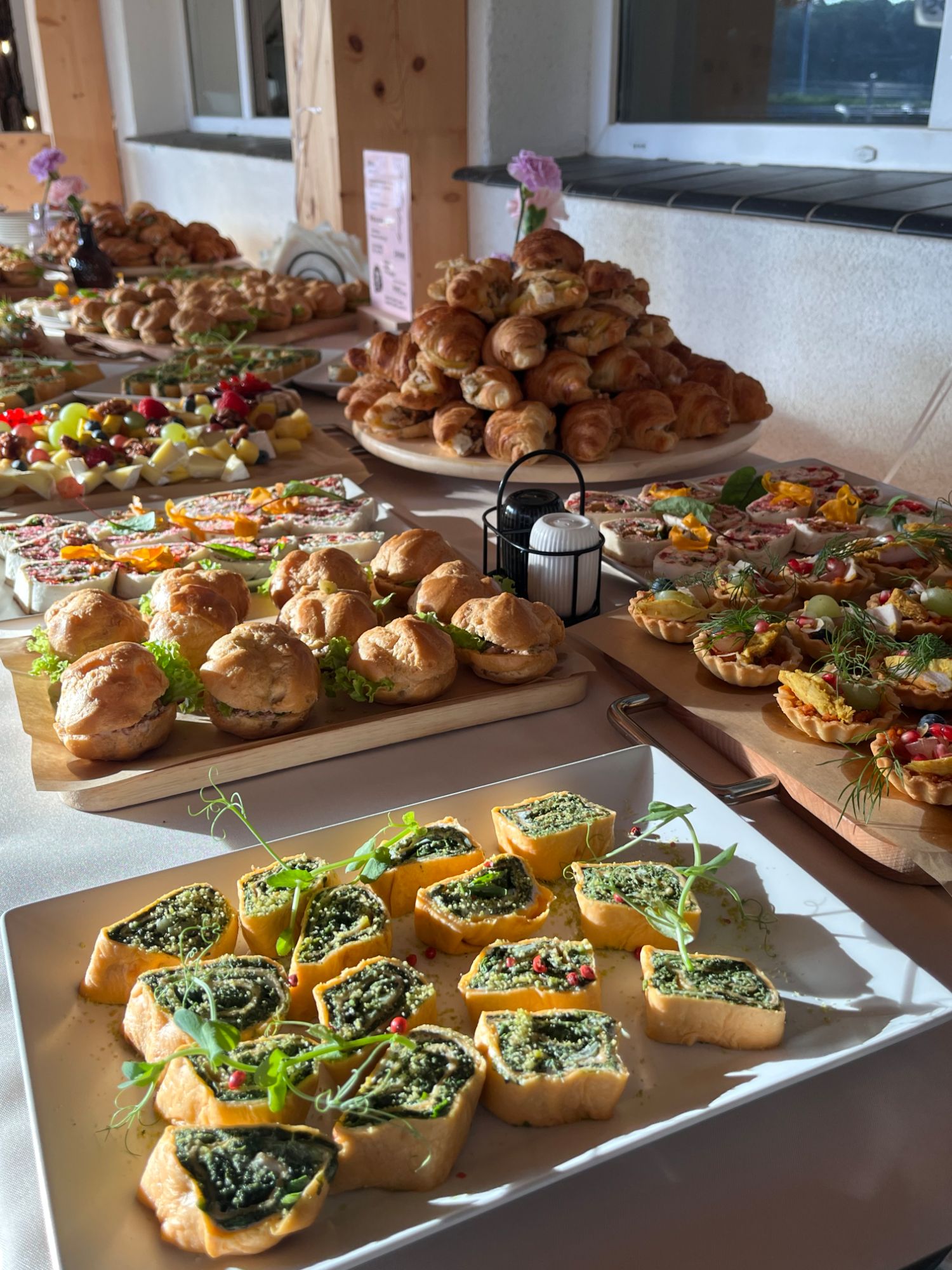 Stół z różnorodnymi przekąskami, w tym ciastkami, sałatkami i tartletkami, w jasnym wnętrzu.