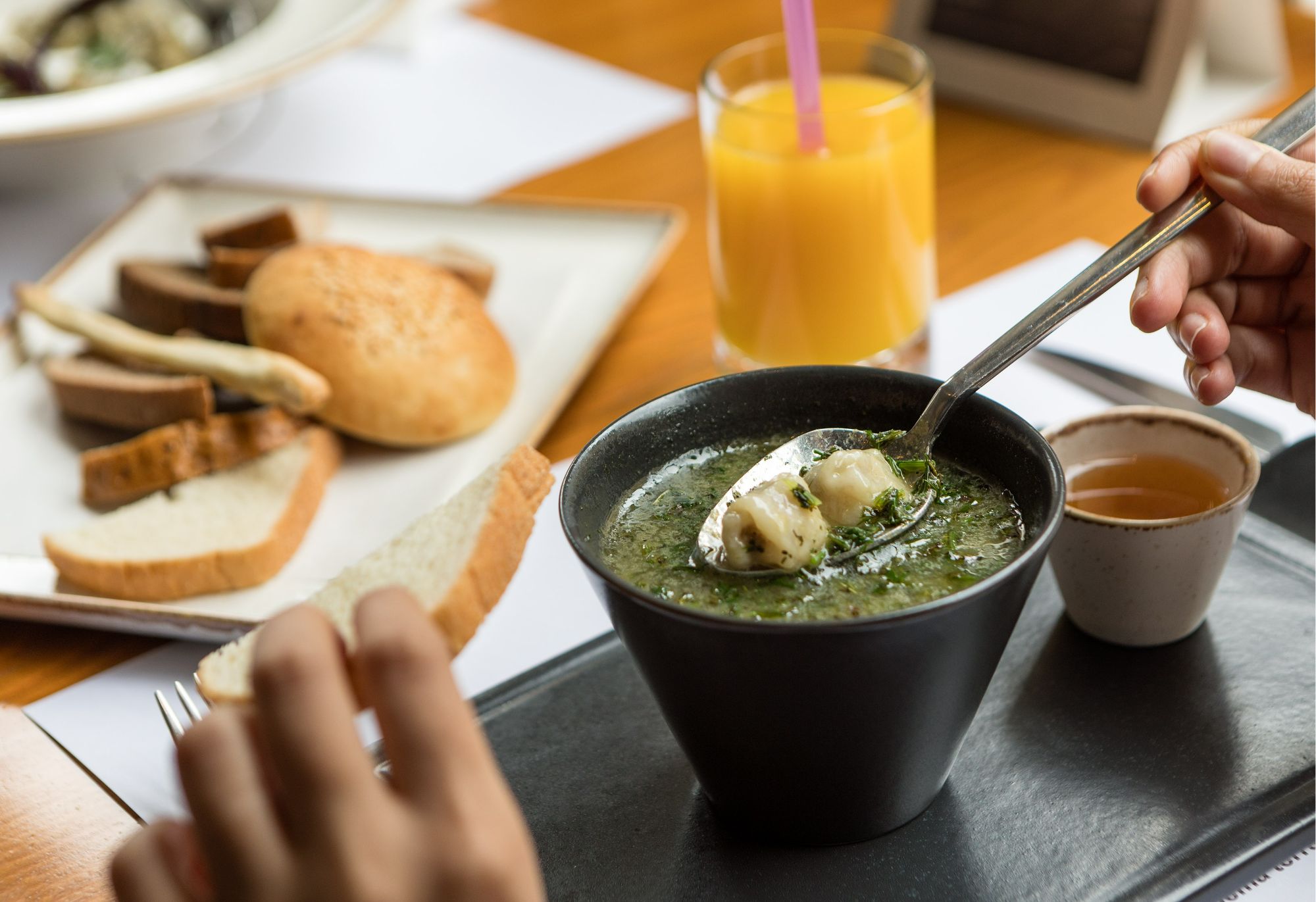 Zupa w czarnej misce, łyżka w zupie, pieczywo i sok pomarańczowy na stole.