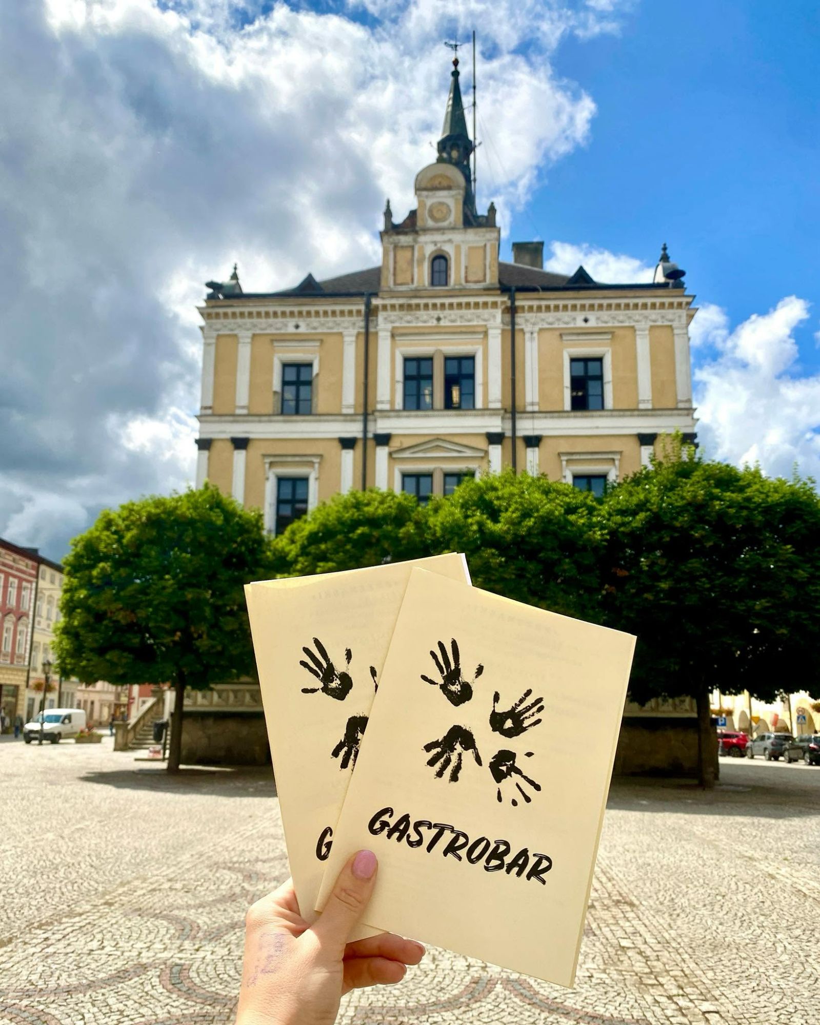 Osoba trzymająca menu „GASTROBAR” przed kolorowym budynkiem z drzewami i chmurami na niebie.