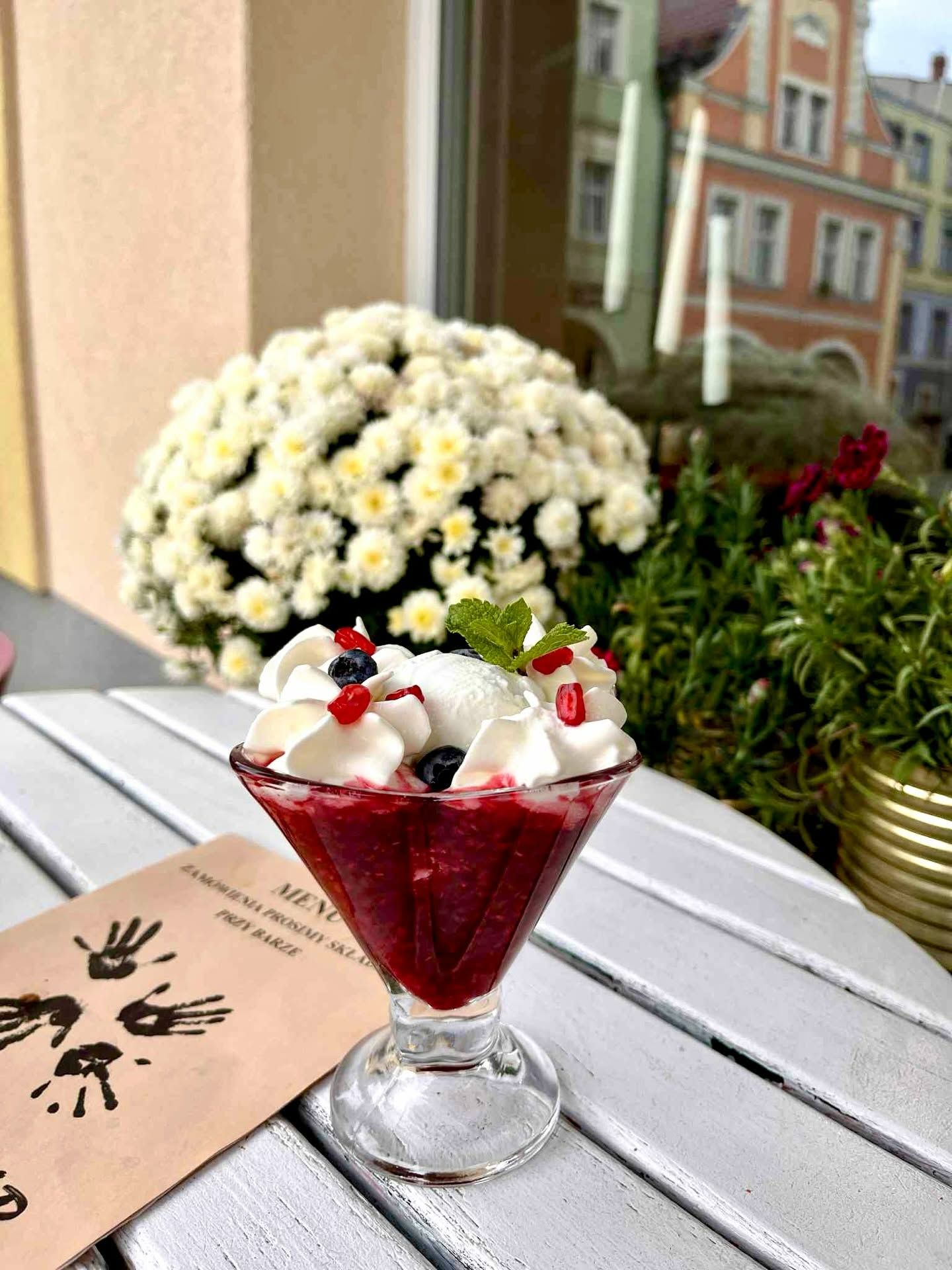 Deser owocowy z bitą śmietaną, dekorowany owocami i miętą, na tle kwitnącej roślinności i menu.