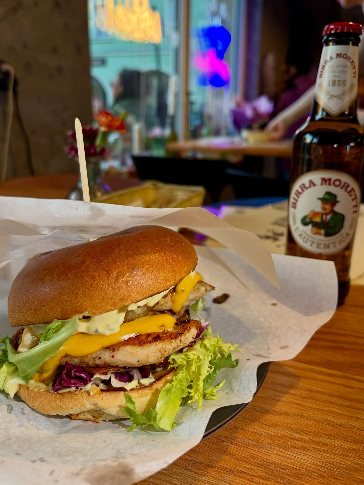 Soczysty burger z kurczakiem, serem, sałatą i sosami, obok butelka piwa i frytki na stole.