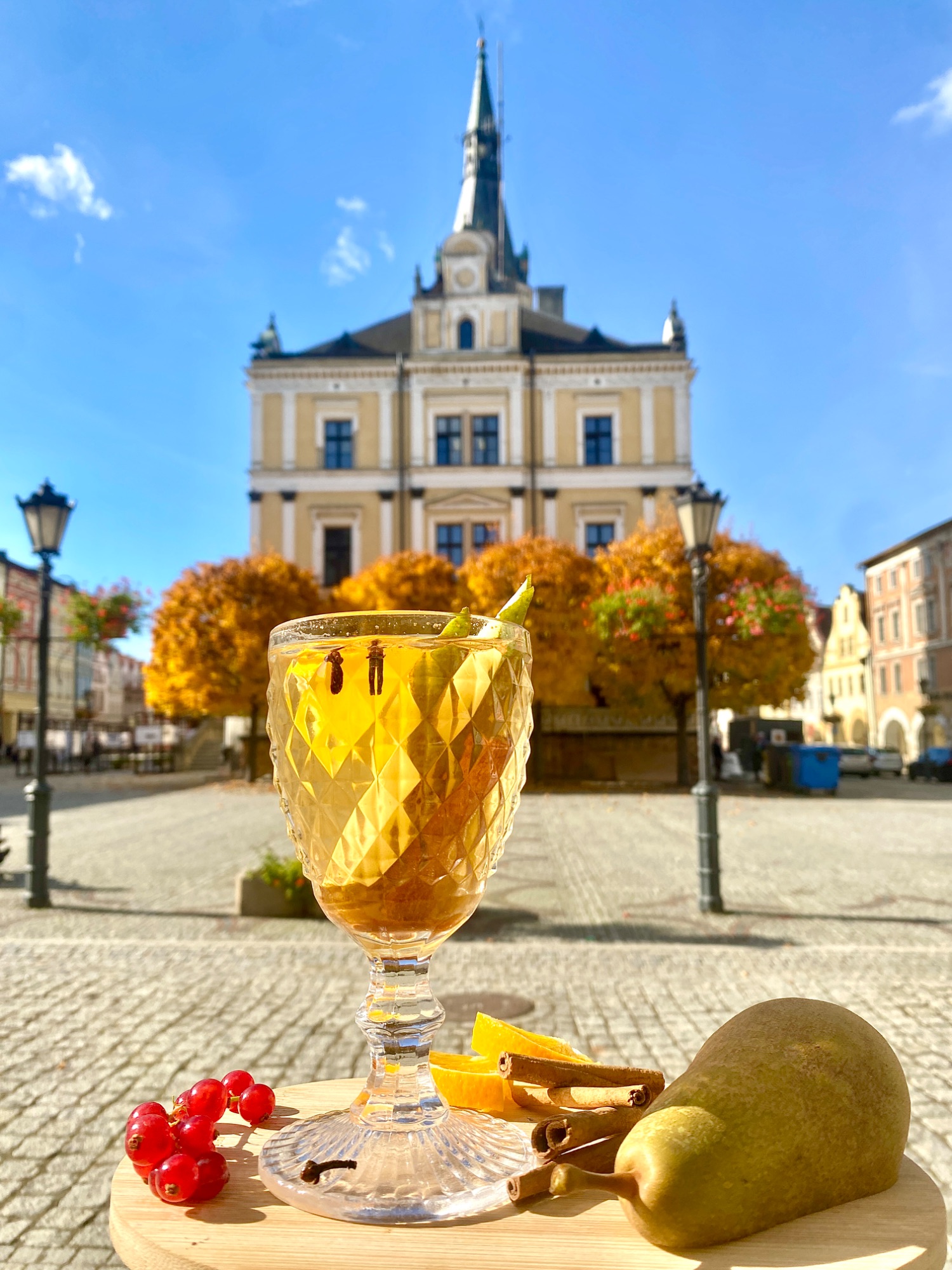 Kieliszek z napojem owocowym na tle zabytkowego budynku, złote liście drzew wokół, słoneczne niebo.