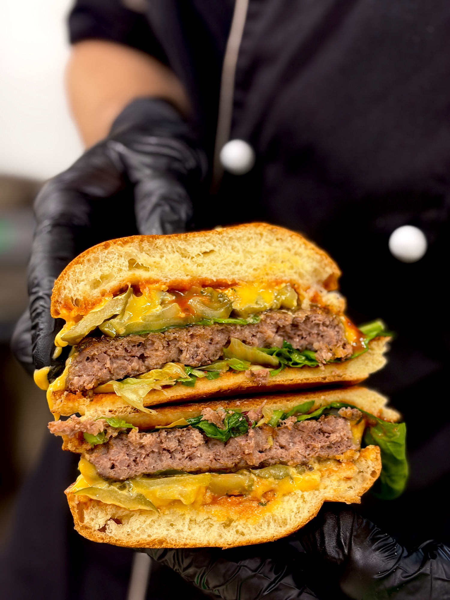 Soczysty burger z wołowiny, serem cheddar, sałatą i piklami, trzymany ręką w czarnych rękawiczkach.