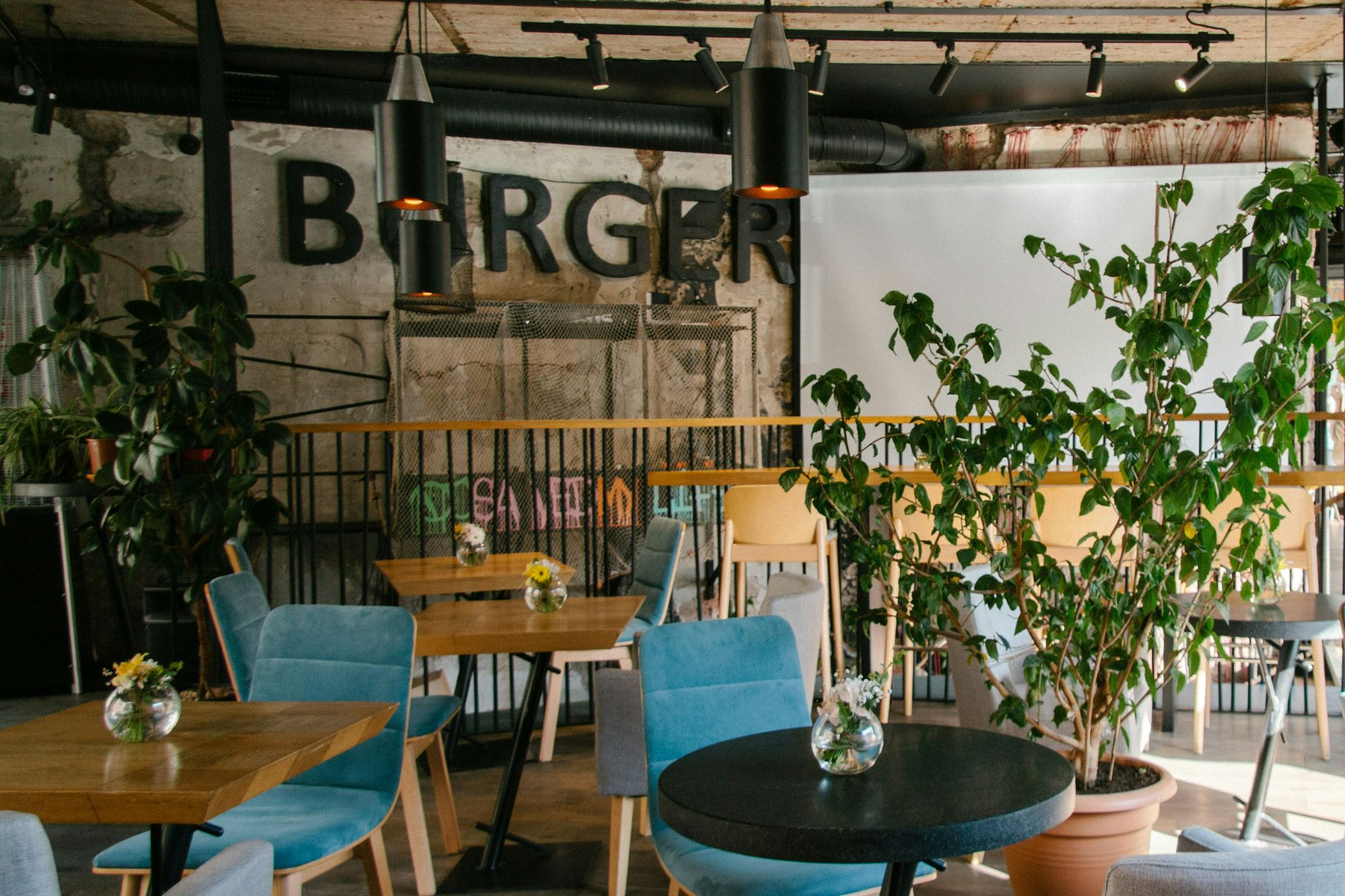 Wnętrze restauracji z niebieskimi krzesłami, drewnianymi stołami, roślinami i dużym napisem "BURGER" na tle.