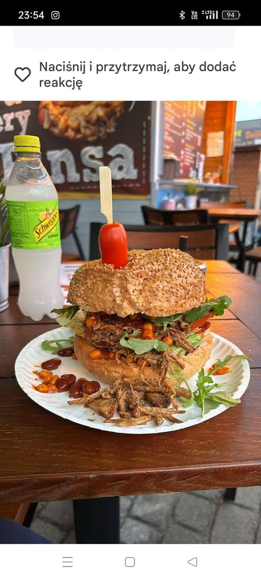 Duży burger z mięsem, warzywami, mayo i pomidorkiem, podany na talerzu obok butelki wody mineralnej.